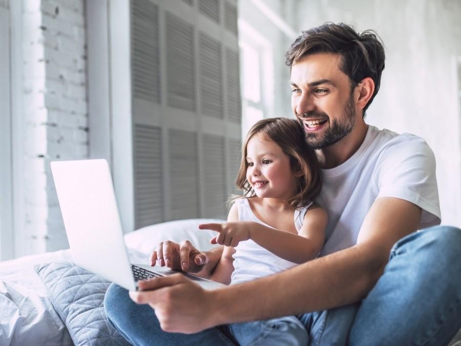 Pflegevater sitzt mit Pflegetochter auf dem Sofa und sie schauen zusammen auf den Bildschirm eines Laptops und lachen. Tagespflegenest, Kita, Kindergarten, Kinderbetreuung, Pflegevater, Pflegemutter, Kindertagespflege.