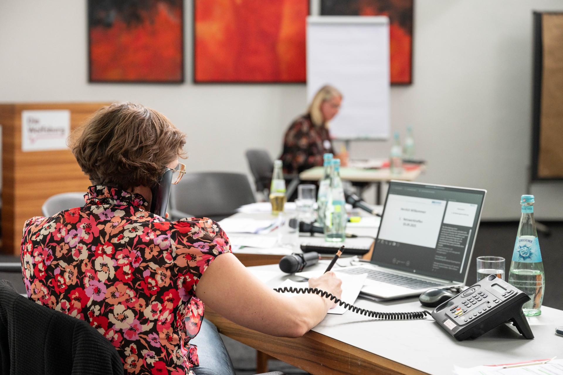 Im Vordergrund sitzt die Teamleitung des Allgemeinen Sozialen Dienstes und telefoniert mit der sich im Hintergrund des Bildes befindenden Schulsozialarbeiterin.
