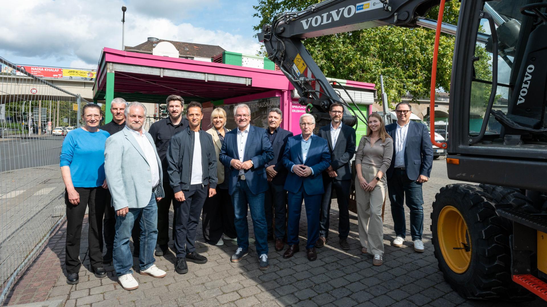 Gruppe von zwölf Personen steht vor einem grün pinken Kiosk. Ein Bagger ist rechts im Bild sichtbar. Der Kiosk ist auf einem gepflasterten Platz.