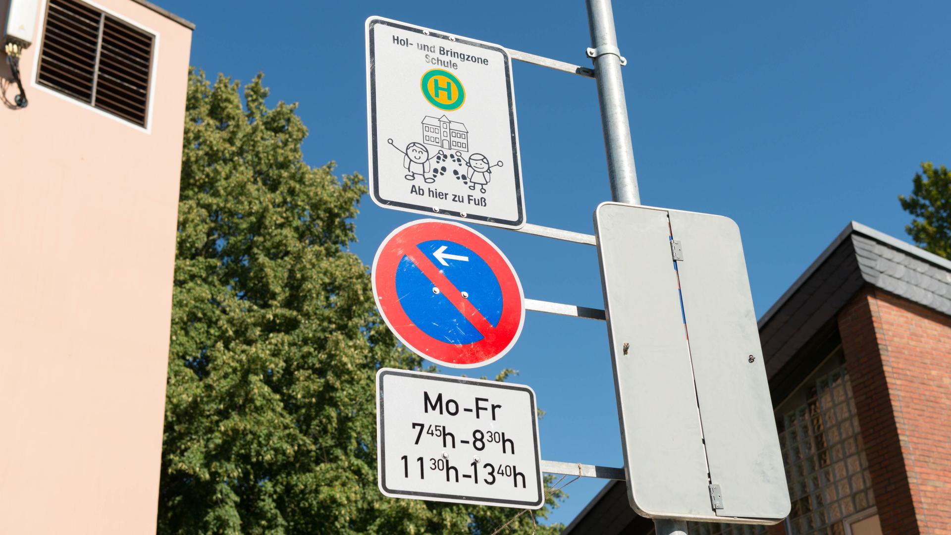 Verkehrsschilder an einem Mast: Haltestelle mit Text 'Hol- und Bringzone Schule Ab hier zu Fuß', ein blau-rotes Parkverbotsschild und weißes Schild.