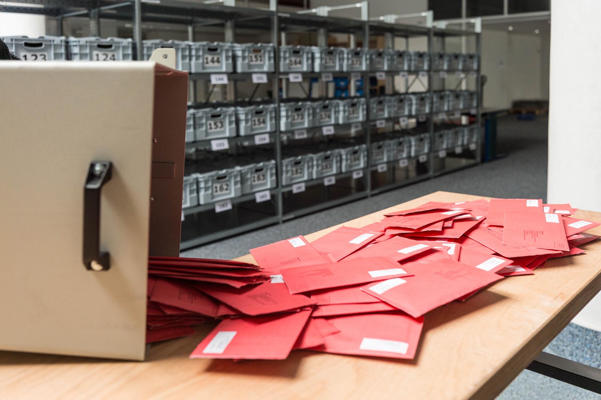 Verschlossene rote Briefwahlumschläge sind aus einer Wahlurne auf einen Tisch geschüttet worden. Im Hintergrund befinden sich Regale mit Metallboxen zur Einlagerung der Stimmzettel aus den jeweiligen Wahlbezirken.