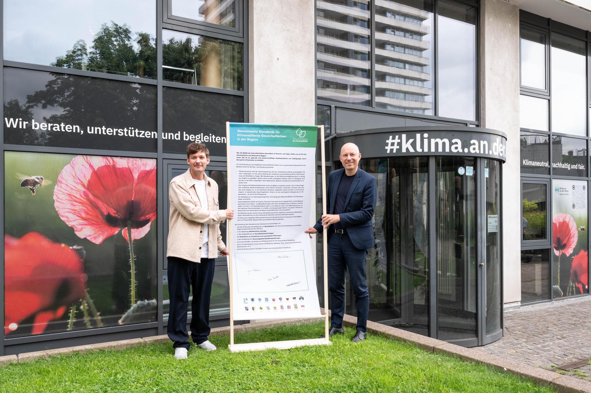Sebastian Mika, Serviceorganisation der Zukunftsinitiative Klima.Werk, sowie Felix Blasch, Beigeordneter für Umwelt, Klima, Bauen, Stadtplanung und Wirtschaftsförderung, stehen vor dem Technischen Rathaus in Mülheim an der Ruhr.