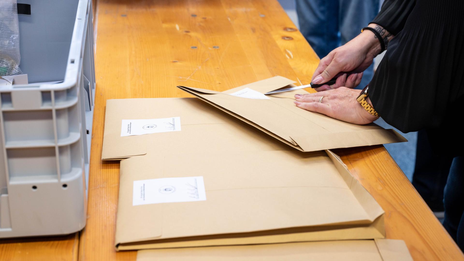 Hände öffnen Versandtaschen mit Siegelmarke auf einem Holztisch, daneben graue Kunststoffbox und Personen im Hintergrund.