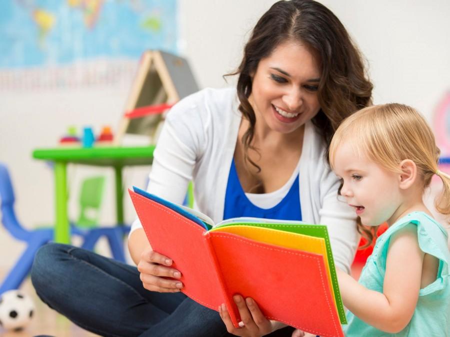 Eine Frau sitzt mit einem kleinem Mädchen in einem Spielzimmer und schaut sich ein Kinderbuch an.