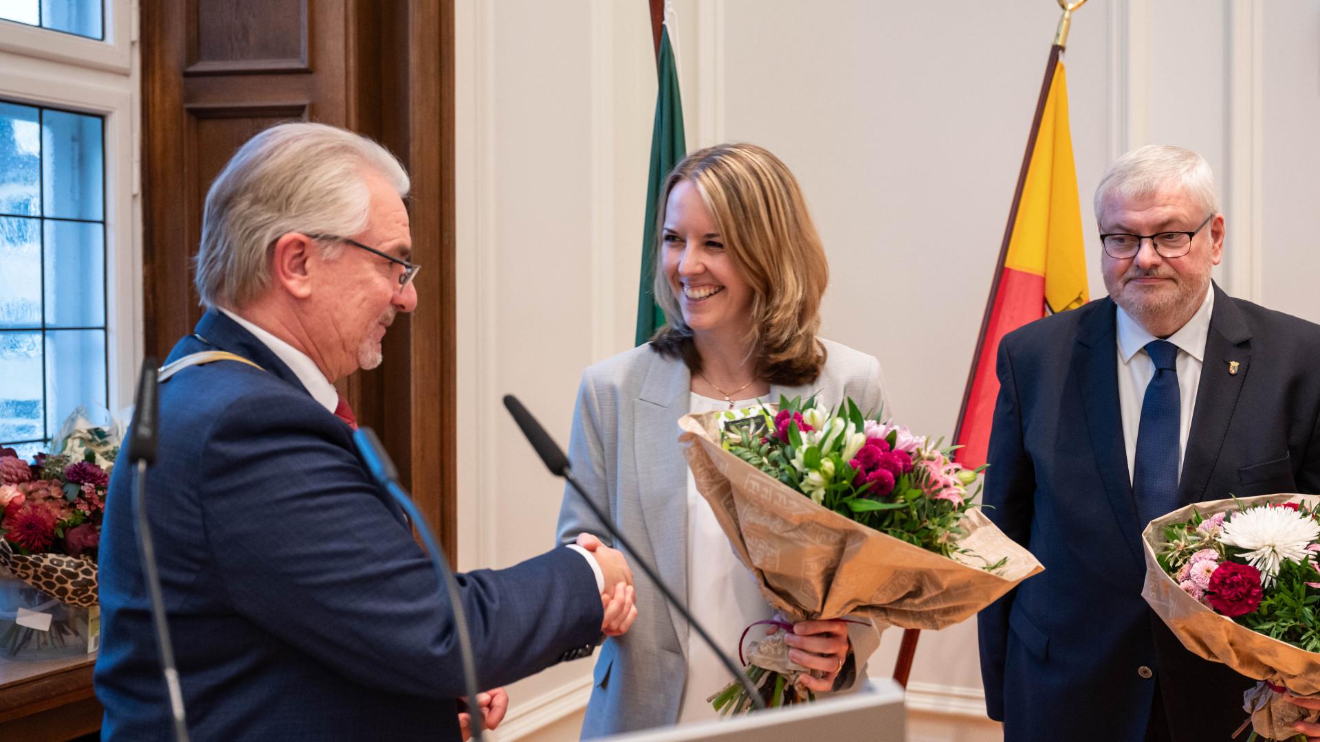 Oberbürgermeister Marc Buchholz der seine ehrenamtliche Stellvertreterin und Stellvertreter gratuliert. Diese haben Blumensträuße in der Hand und stehen vor der Mülheim Flagge.