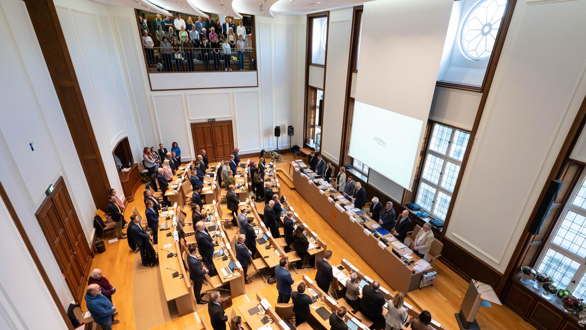 Rat der Stadt tagt im Ratssaal mit mehreren Reihen von Tischen, an denen zahlreiche Personen stehen. Große Fenster und ein Balkon mit weiteren Zuschauern sind sichtbar.