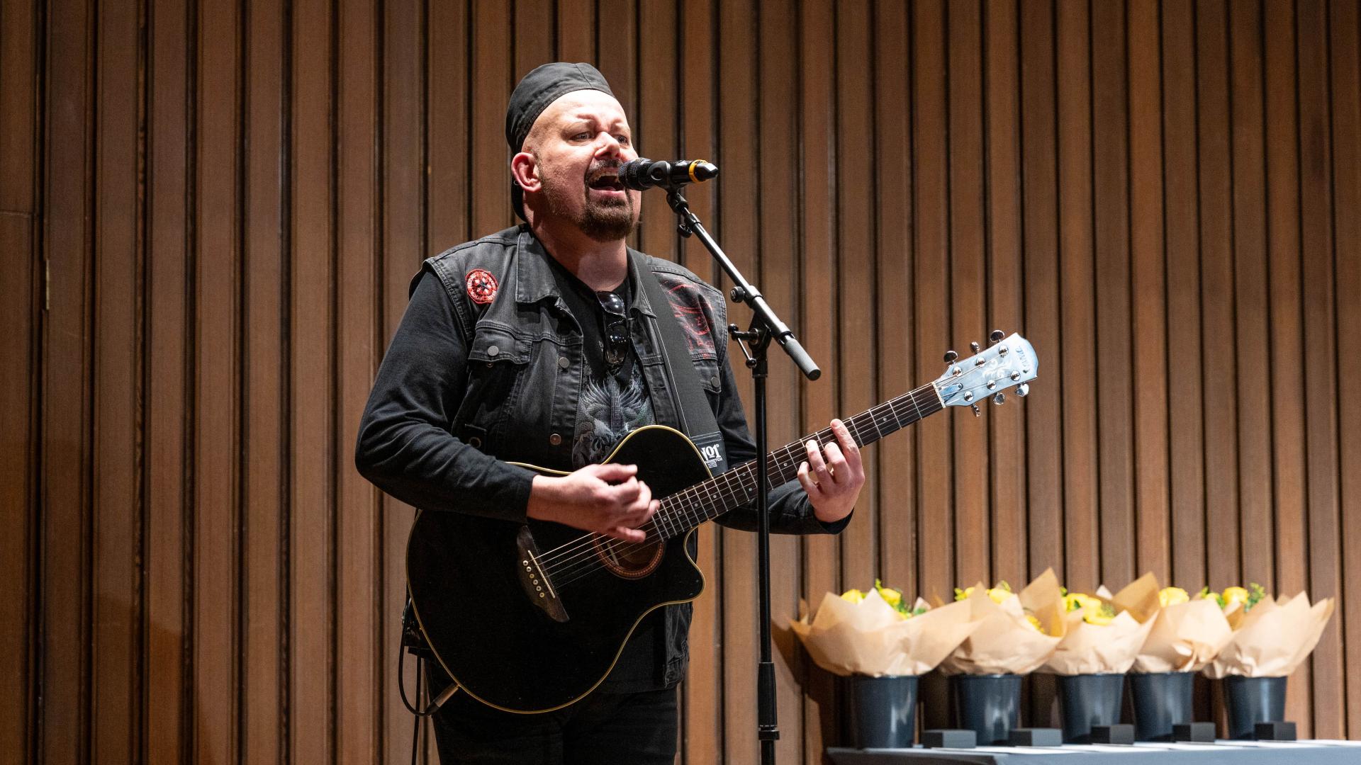Ein Mann mit einer Gitarre steht vor einer holzvertäfelten Wand und singt in ein Mikrofon. Im Hintergrund sind mehrere Blumensträuße zu sehen.