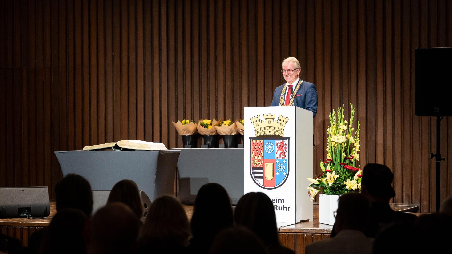 Der Oberbürgermeister hält auf der Bühne an einem Stehpult eine Rede. Im Vordergrund sind die Köpfe des Publikums zu sehen.