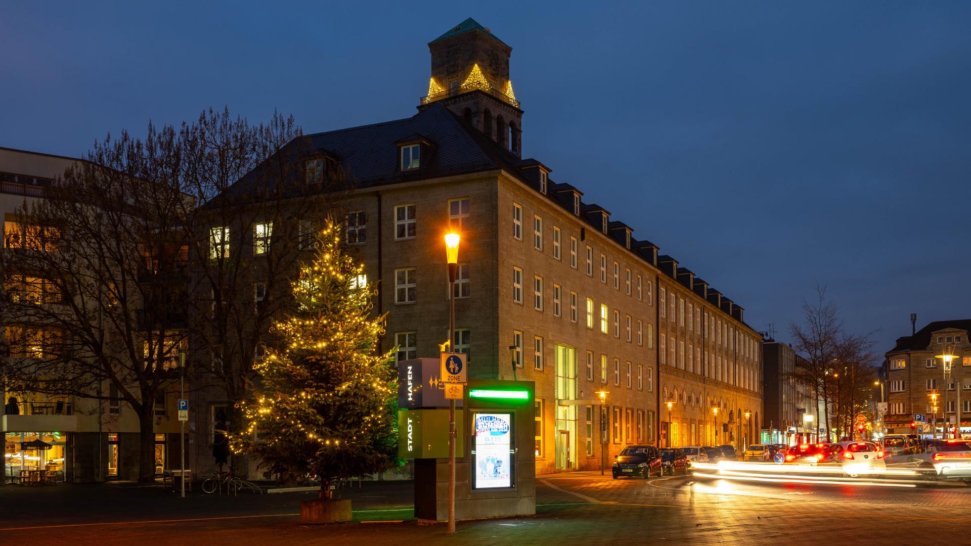 Weihnachtsbaum mit gelben Lichtern vor dem Rathaus in Mülheim an der Ruhr bei Abenddämmerung. Im Hintergrund der Rathausturm mit Weihnachtsbeleuchtung.