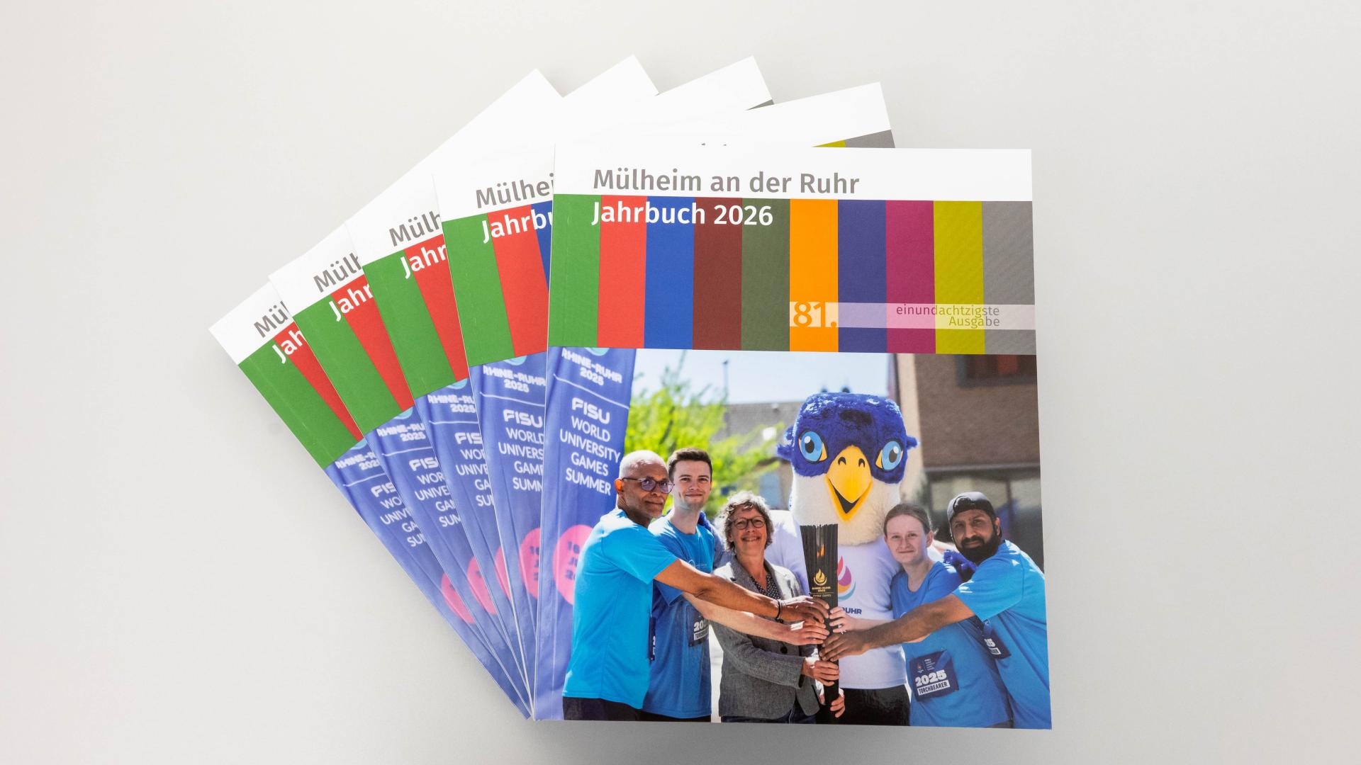 Mehrere Jahrbücher 2026 von Mülheim an der Ruhr liegen fächerförmig, vorne ein Jahrbuch mit vier Personen in blauen T-Shirts und einer Person in grauem Anzug, die gemeinsam eine Fackel halten, daneben eine Person in Vogelmaskottchen-Kostüm.