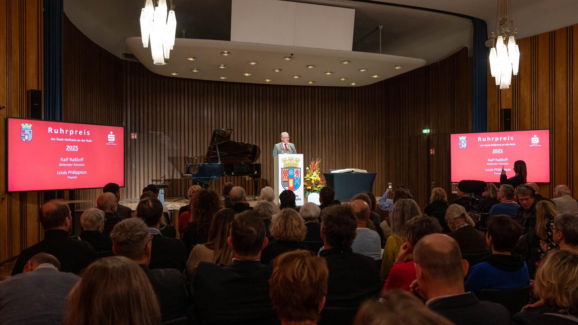 Oberbürgermeister Marc Buchholz steht am Podium mit Wappen, Publikum sitzt in Saal mit zwei großen Bildschirmen, die 'Ruhrpreis 2025' anzeigen.