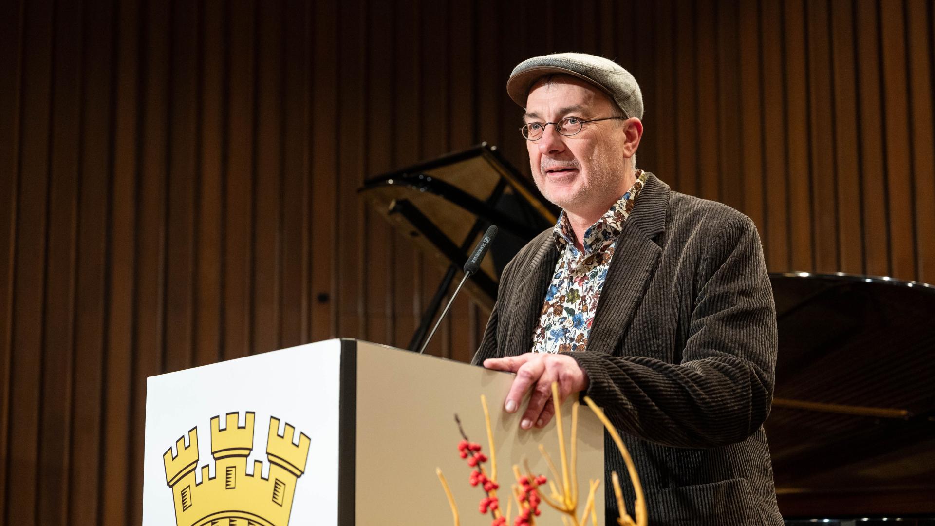 Ralf Raßloff in braunem Cordjackett steht hinter einem Rednerpult mit einem Wappen, im Hintergrund ein schwarzes Klavier.