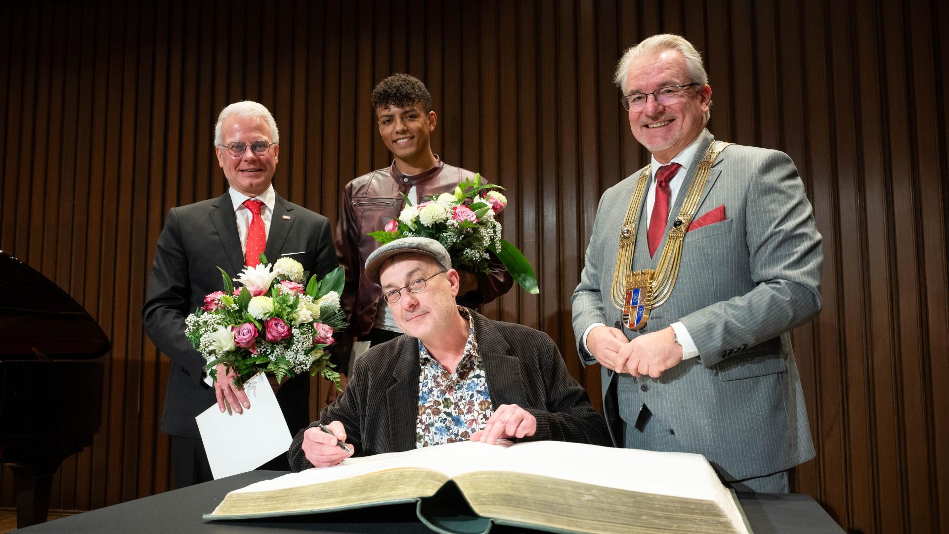 Vier Personen in formeller Kleidung, eine sitzt und trägt sich ins Goldene Buch der Stadt ein, zwei halten Blumensträuße, der Oberbürgermeister trägt eine goldene Amtskette.