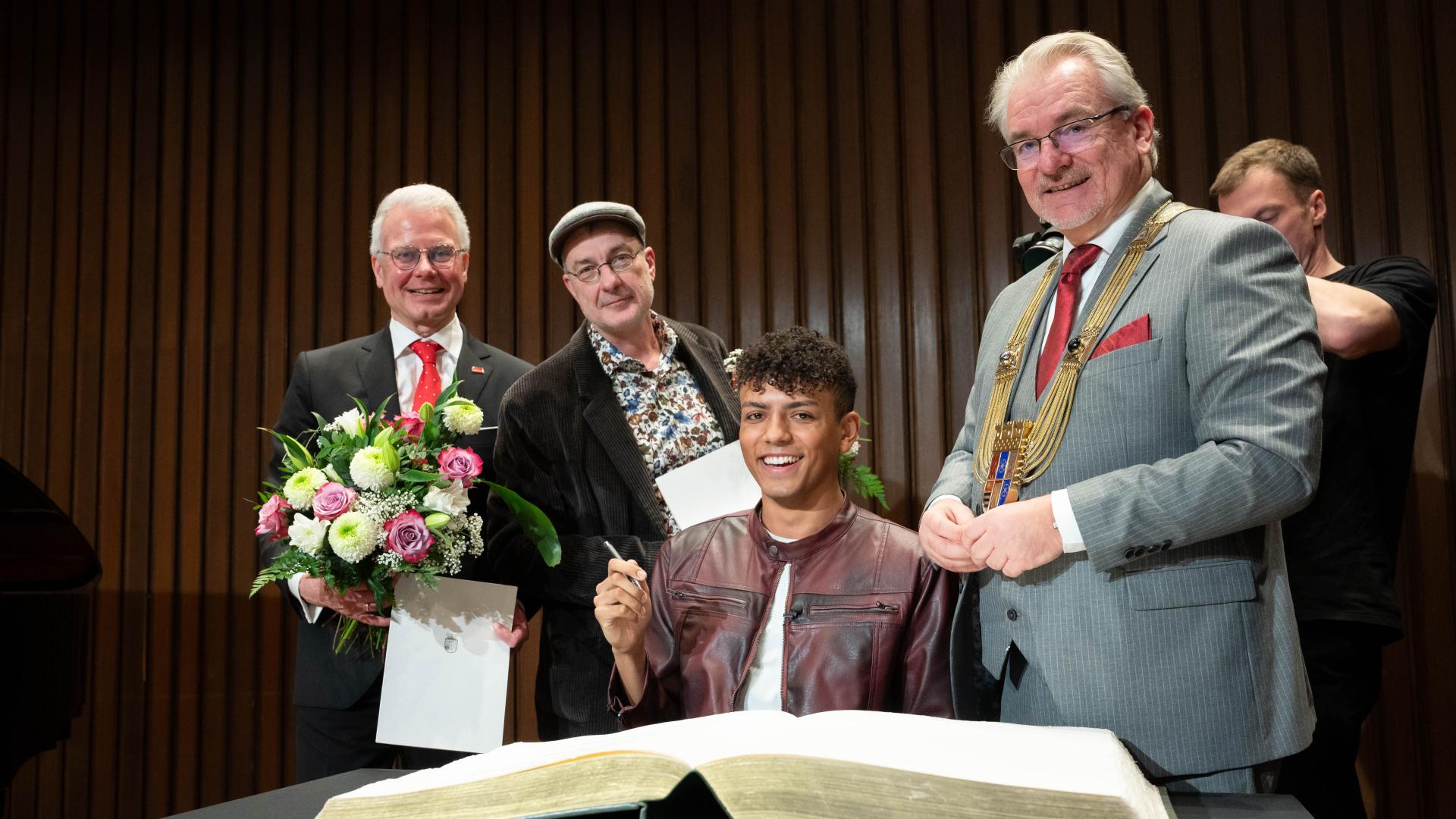Vier Personen in formeller Kleidung, eine sitzt und trägt sich ins Goldene Buch der Stadt ein, zwei halten Blumensträuße, der Oberbürgermeister trägt eine goldene Amtskette.
