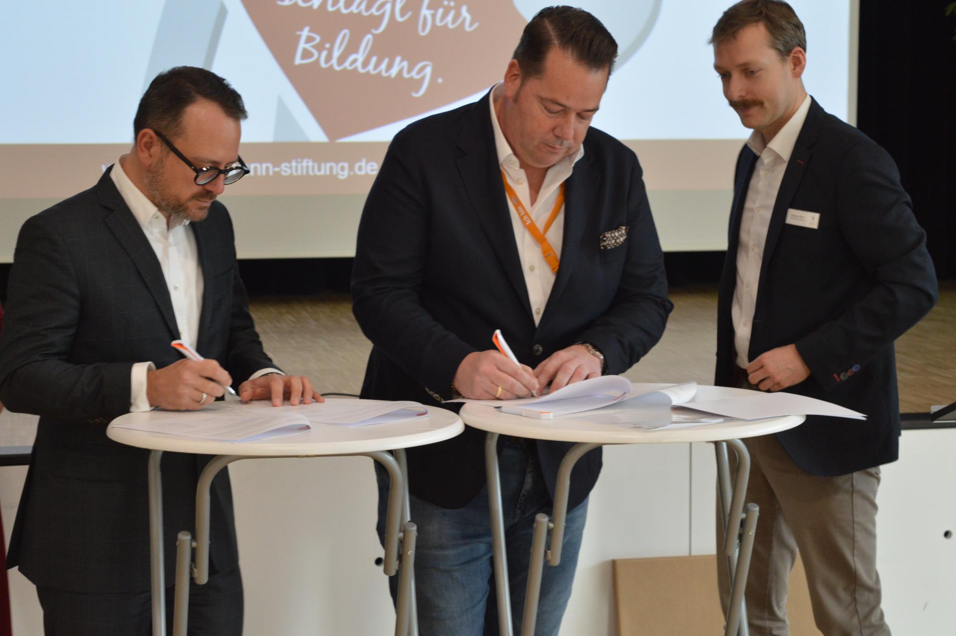Zwei Personen an Stehtischen unterschreiben die Kooperationsvereinbarung. Im Hintergrund ist eine dritte Person zu sehen.