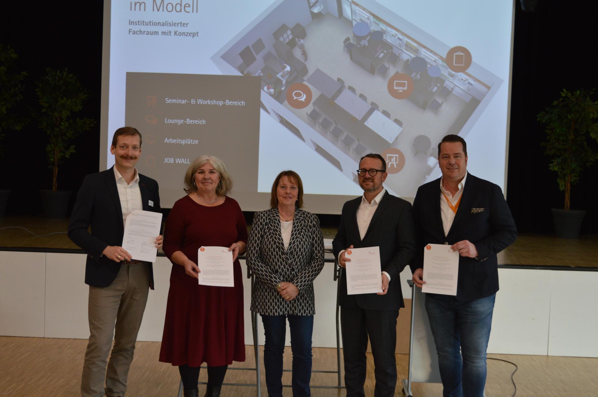 Fünf Beteiligte stehen auf einer Bühne und halten die Kooperationsvereinbarung in die Kamera. Im Hintergrund ist per Beamter eine Projektion an die Wand geworfen.
