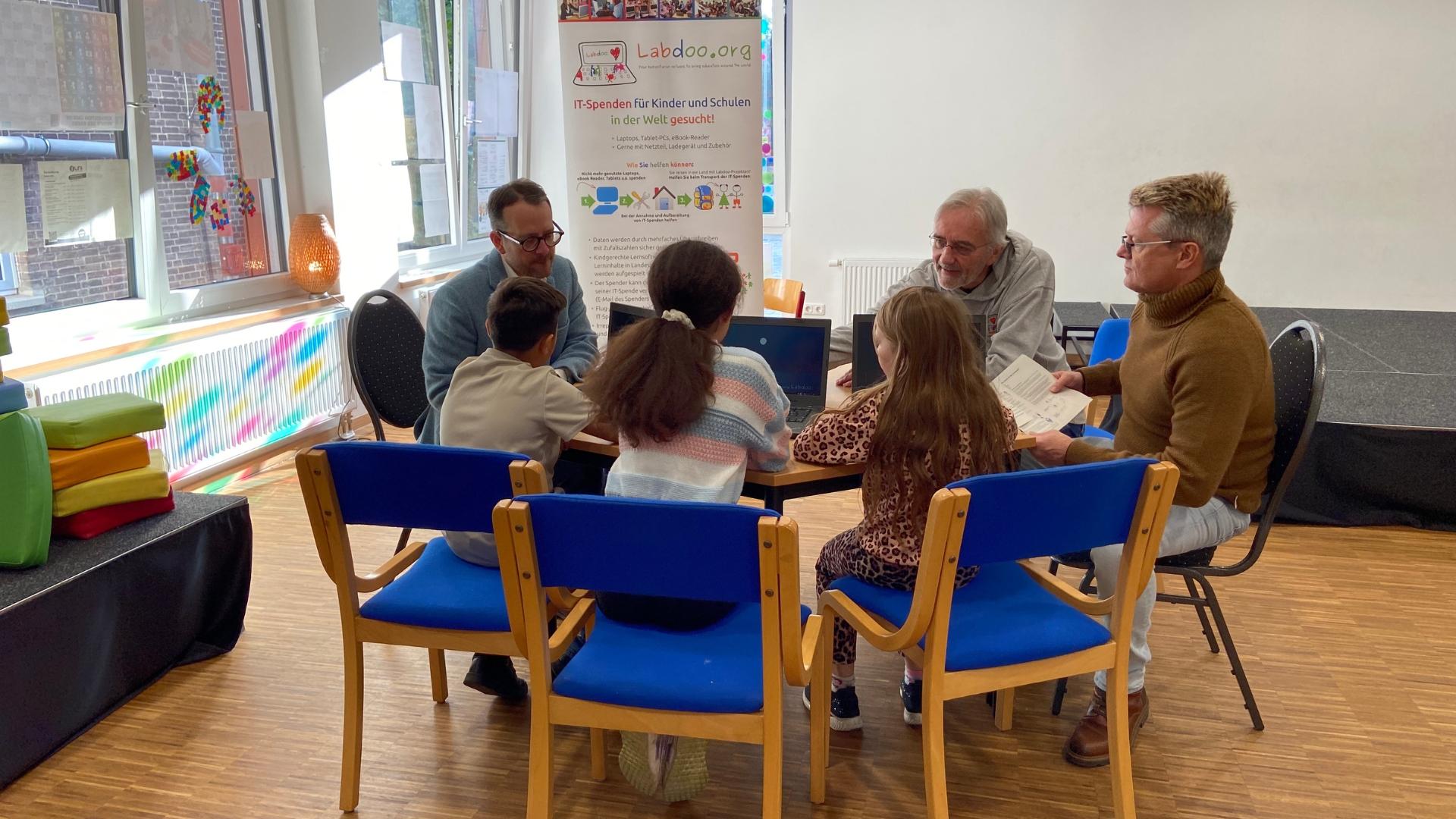 Drei Erwachsene sitzen zusammen mit drei Kindern an einem Tisch in einem Raum an der Brüder Grimm Schule in Mülheim.