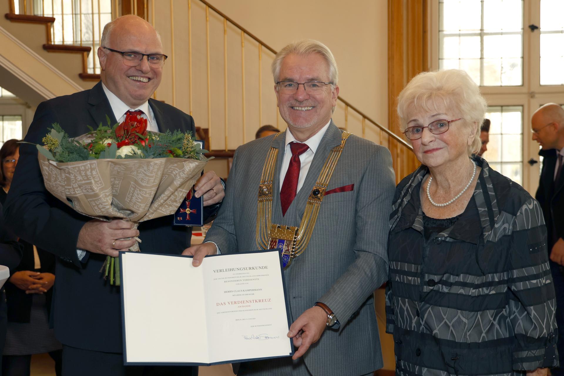 Ein Mann hält einen Blumenstrauß und zeigt das Verdienstkreuz in die Kamera. Neben ihm steht der Oberbürgermeister mit der Verleihungsurkunde und daneben die Frau des Ausgezichneten.