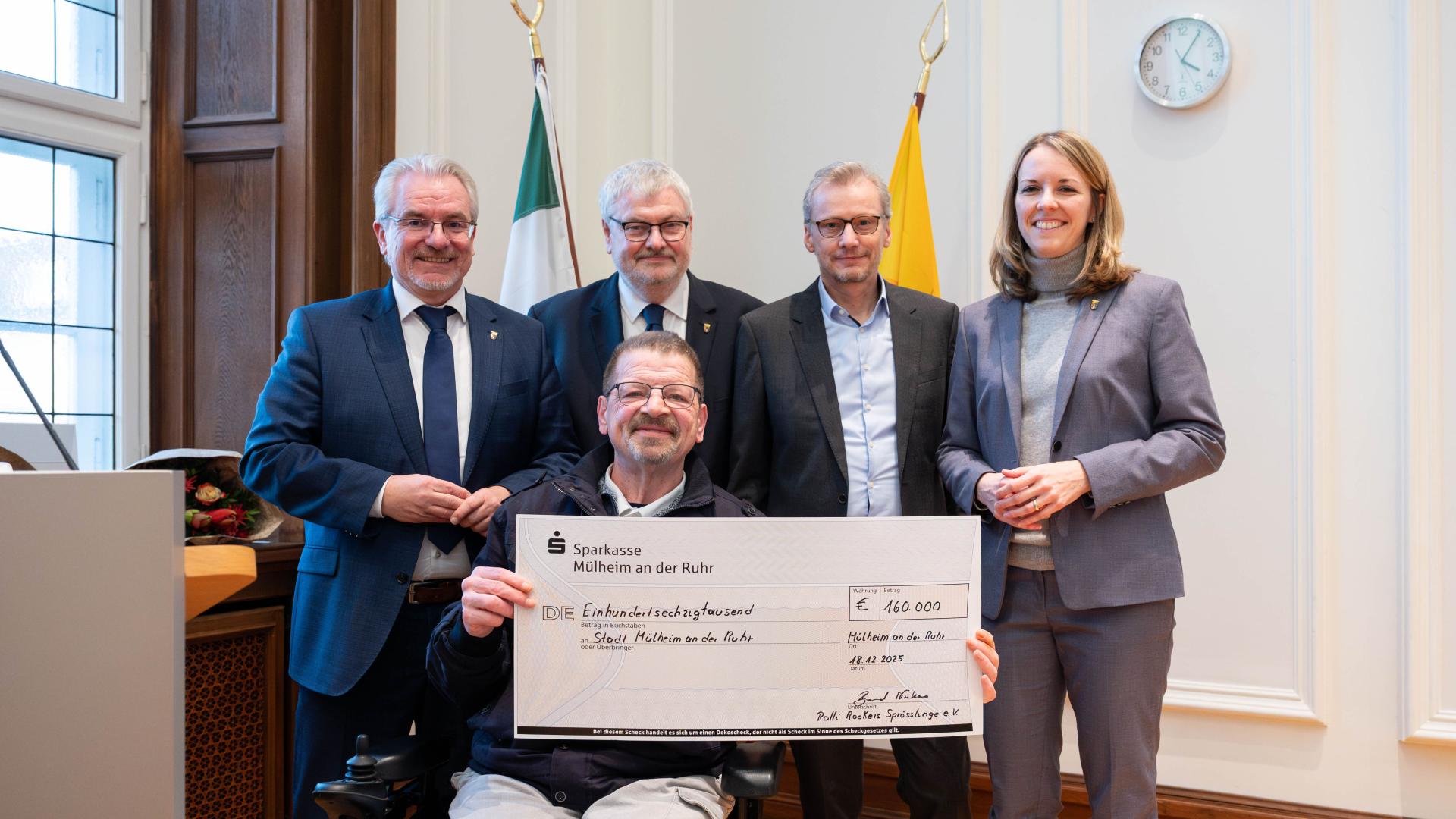 Vier Personen stehen hinter einer sitzenden Person, die einen großen symbolischen Scheck erhalten hat (Rolli Rockers). Ratssaal