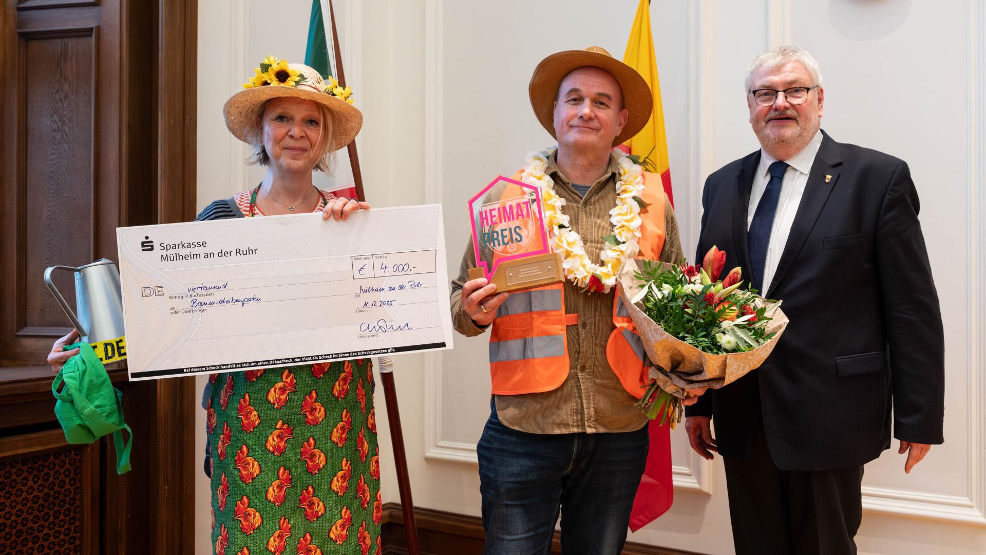 Drei Personen stehen im Ratssaal, eine hält einen großen Scheck, eine weitere eine Auszeichnung und einen Blumenstrauß.