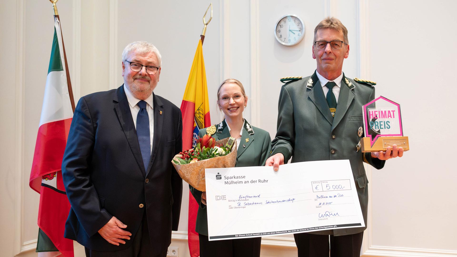 Drei Personen in formeller Kleidung, eine hält einen großen Scheck über 6.500 Euro, eine andere einen Blumenstrauß und eine Trophäe mit der Aufschrift 'Heimat Preis'.