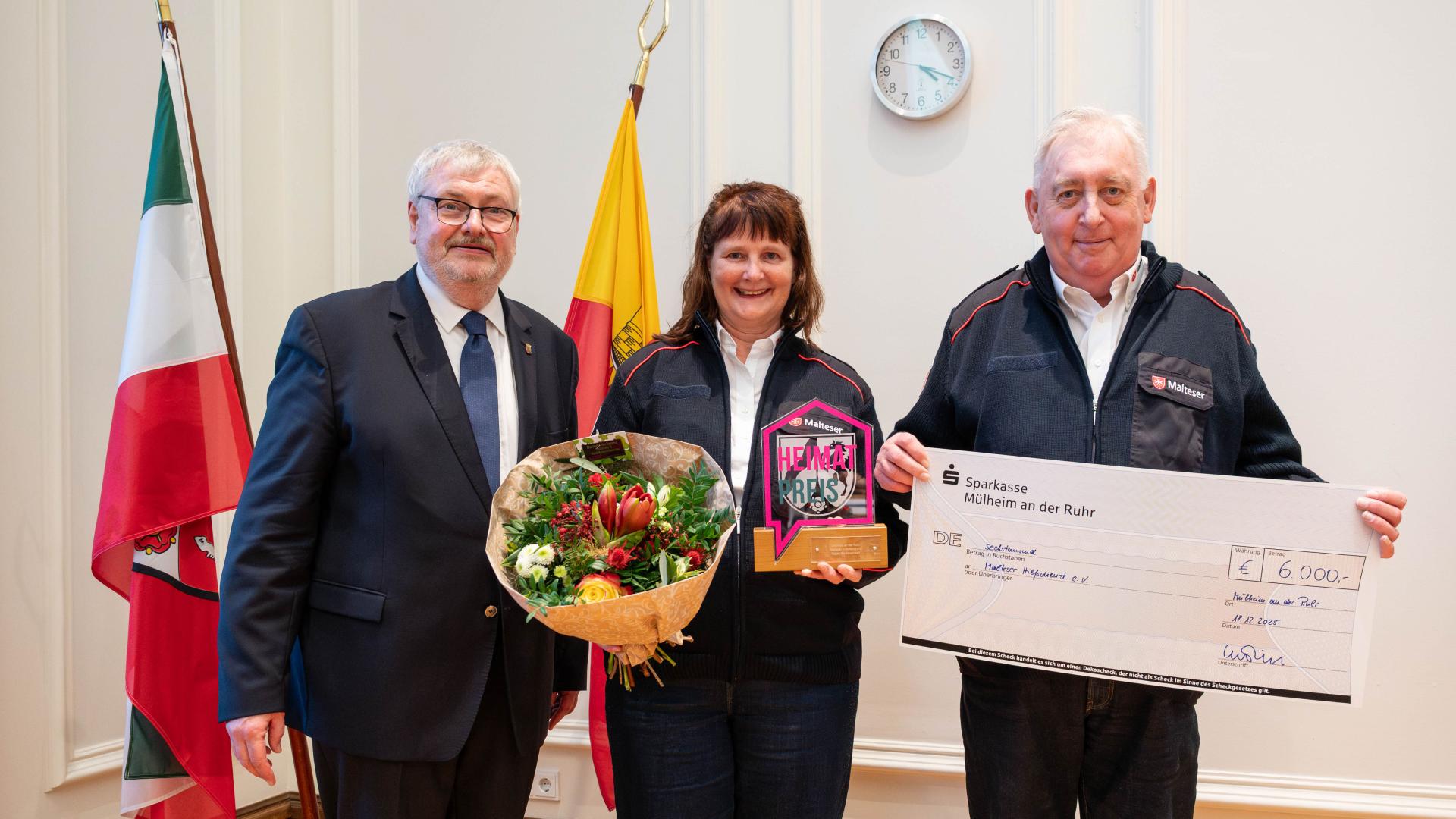 Drei Personen stehen nebeneinander, eine hält einen Blumenstrauß und eine Auszeichnung, eine weitere einen großen Scheck über 6.000 Euro.
