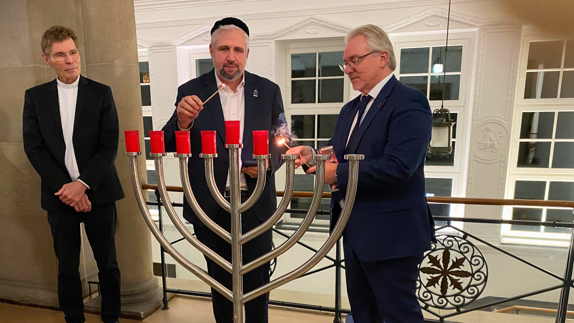 Drei Männer stehen hinter einem großen neunarmigen Chanukka-Kerzenleuchter. Zwei von ihnen, der Rabbiner und der Oberbürgermeister, zünden mit langen Streichhölzern fünf Kerzen am Leuchter an.