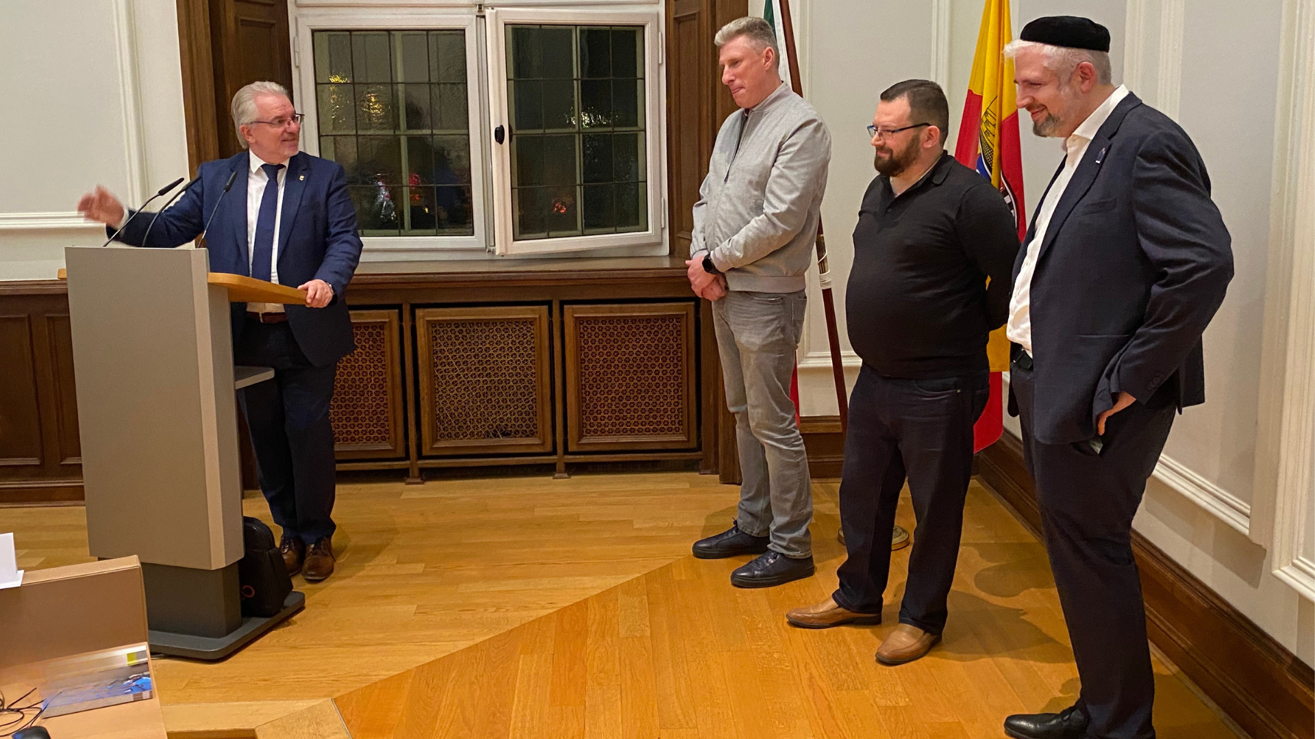Der Oberberbürgermeister steht im Ratssaal am Rednerpult und begrüßt drei Mitglieder der Jüdischen Gemeinde, die rechts neben ihm stehen.