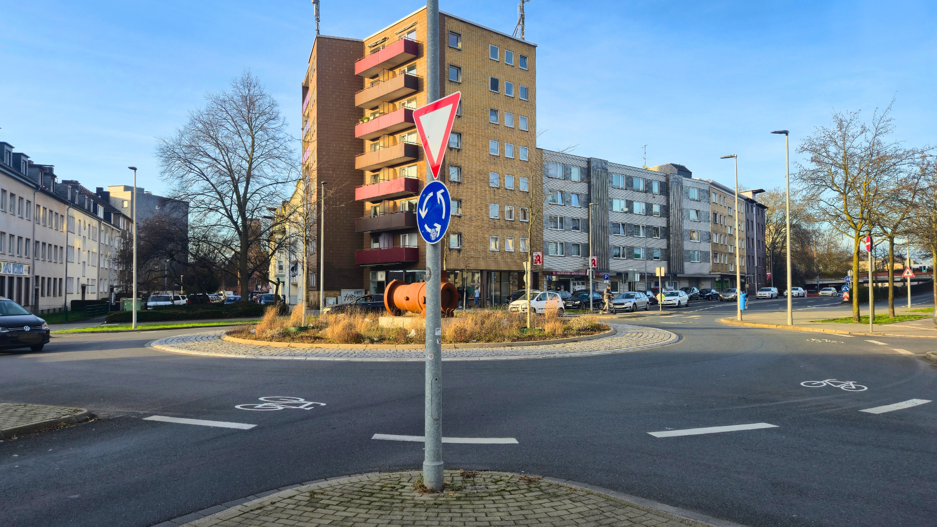 Abgebildet ist ein Kreisverkehr. Im Hintergrund befinden sich Gebäude. Auf der Fahrbahn des Kreisels sind Piktogramme aufgebracht. Es handelt sich um weiße Fahrräder.