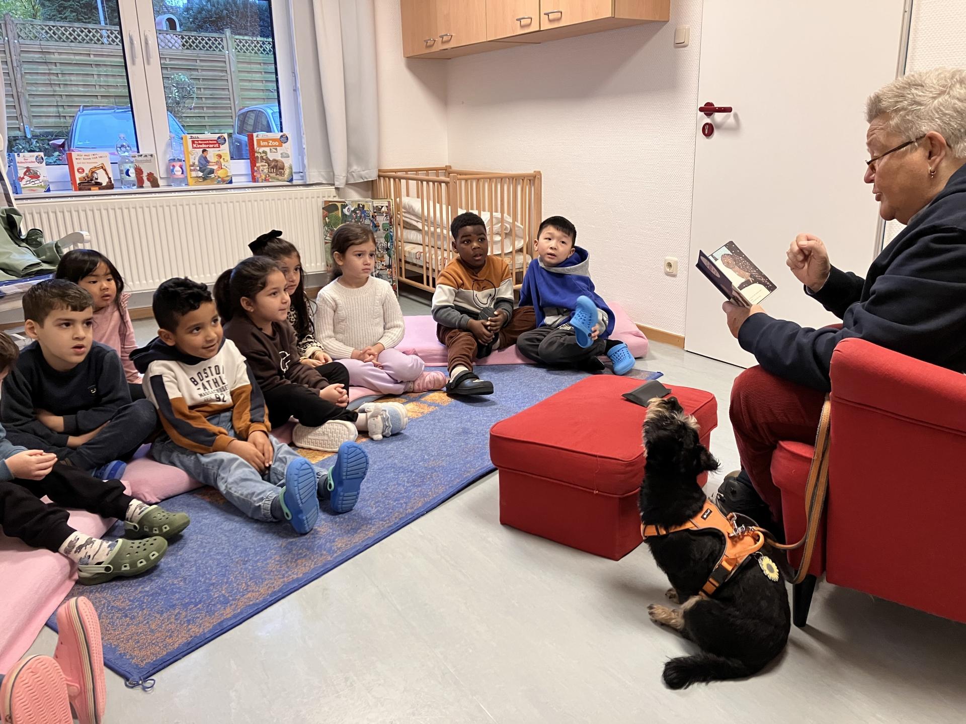 Eine Frau sitzt in einem roten Sessel, neben ihr ein Hund und liest Kindern vor, die auf dem Boden sitzen.