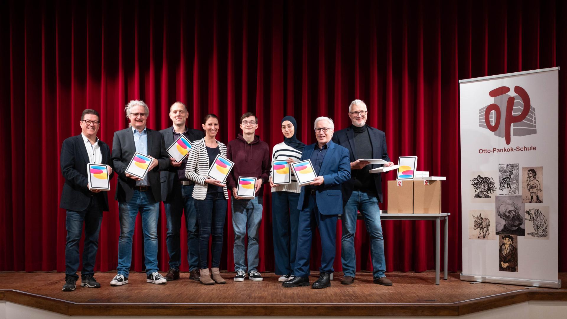 Gruppe von acht Personen auf Bühne vor rotem Vorhang hält iPads in der Hand neben Banner der Otto-Pankok-Schule.