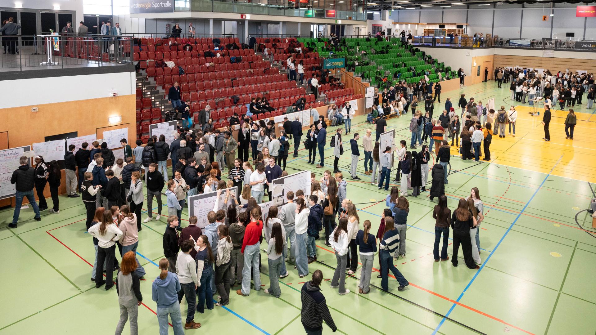 Große Gruppe von Menschen in einer Sporthalle, die an mehreren Stellwänden und Tischen stehen und sich unterhalten. Jugendbezirksforum, Westenergie Sporthalle