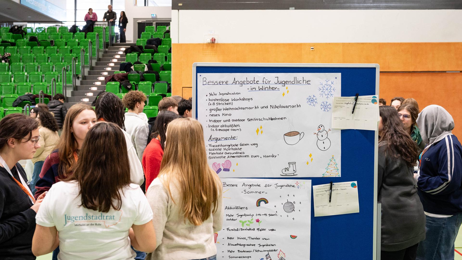 Gruppe Jugendlicher steht vor einer Pinnwand mit handgeschriebenen Plakaten in einer Halle mit grünen Sitzreihen und einer Empore mit zwei Personen. Jugendbezirksforum, Westenergie Sporthalle