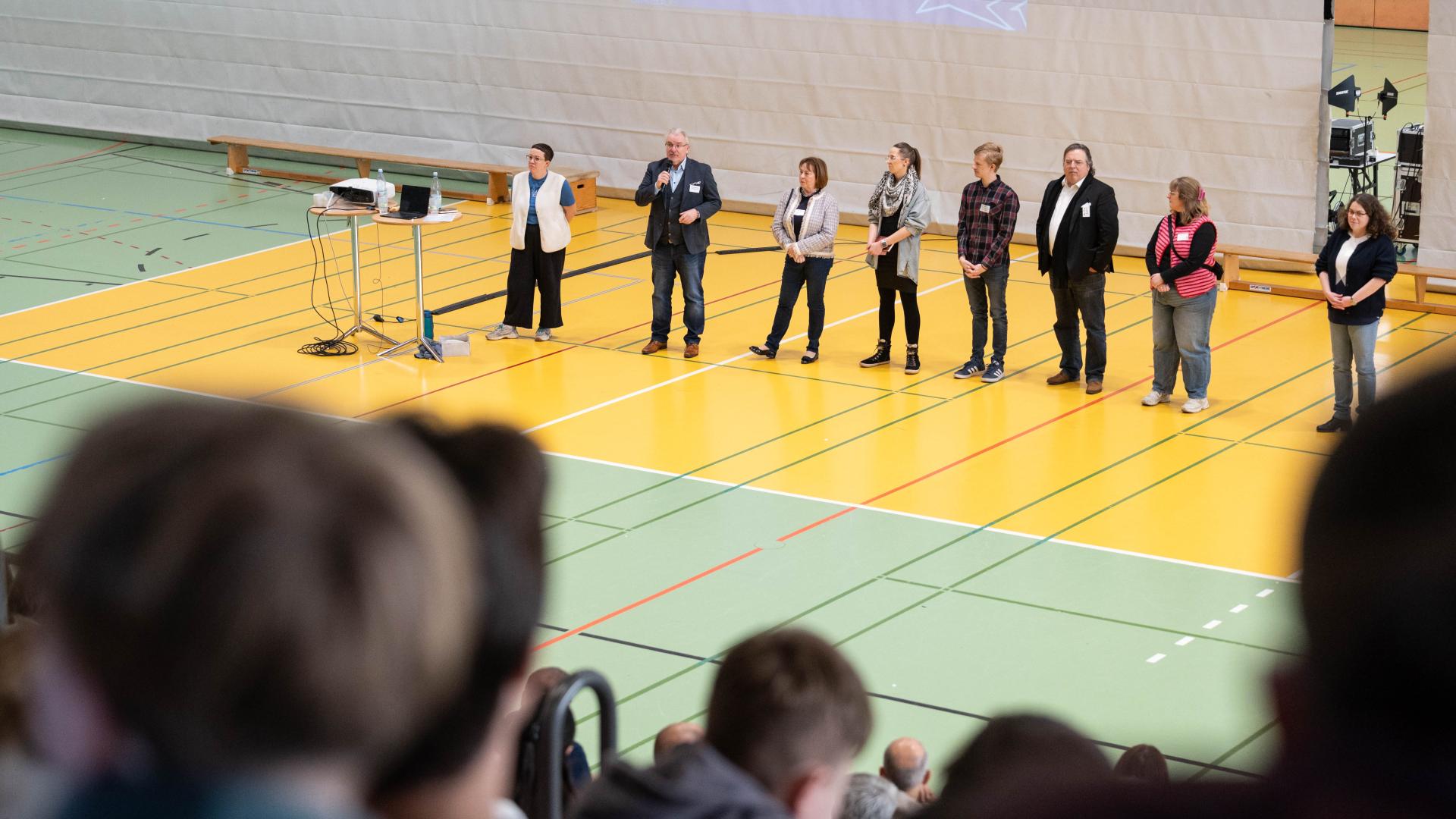 Sieben Personen stehen in einer Reihe auf dem Boden einer Sporthalle, eine Person spricht in ein Mikrofon, im Vordergrund sind verschwommene Köpfe von Zuschauern zu sehen. Jugendbezirksforum, Westenergie Sporthalle