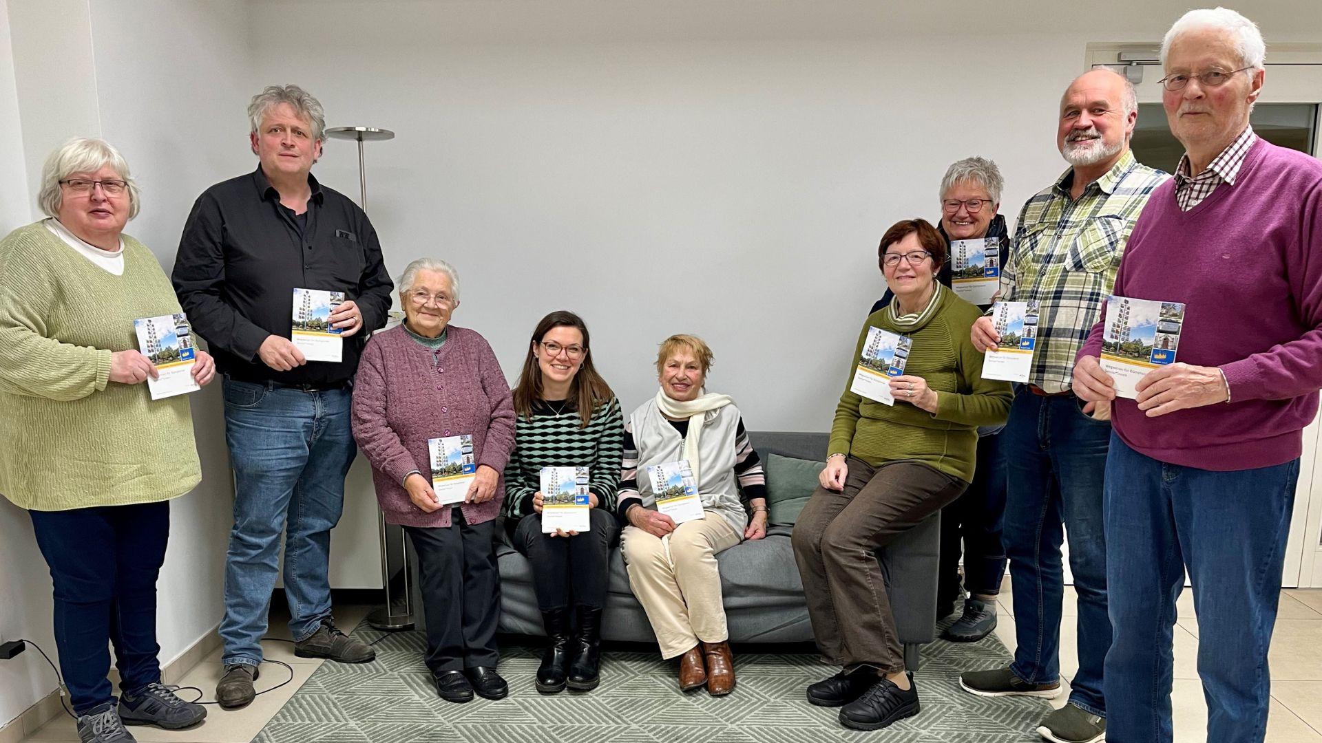 Die Mitglieder der Arbeitsgruppe Dümpten aus dem Netzwerk der Generationen haben sich für ein Gruppenbild aufgestellt. In der Mitte sitzen einige Personen auf einem Sofa, links und rechts daneben stehen weitere Personen und halten den neuen Wegweiser in die Kamera.