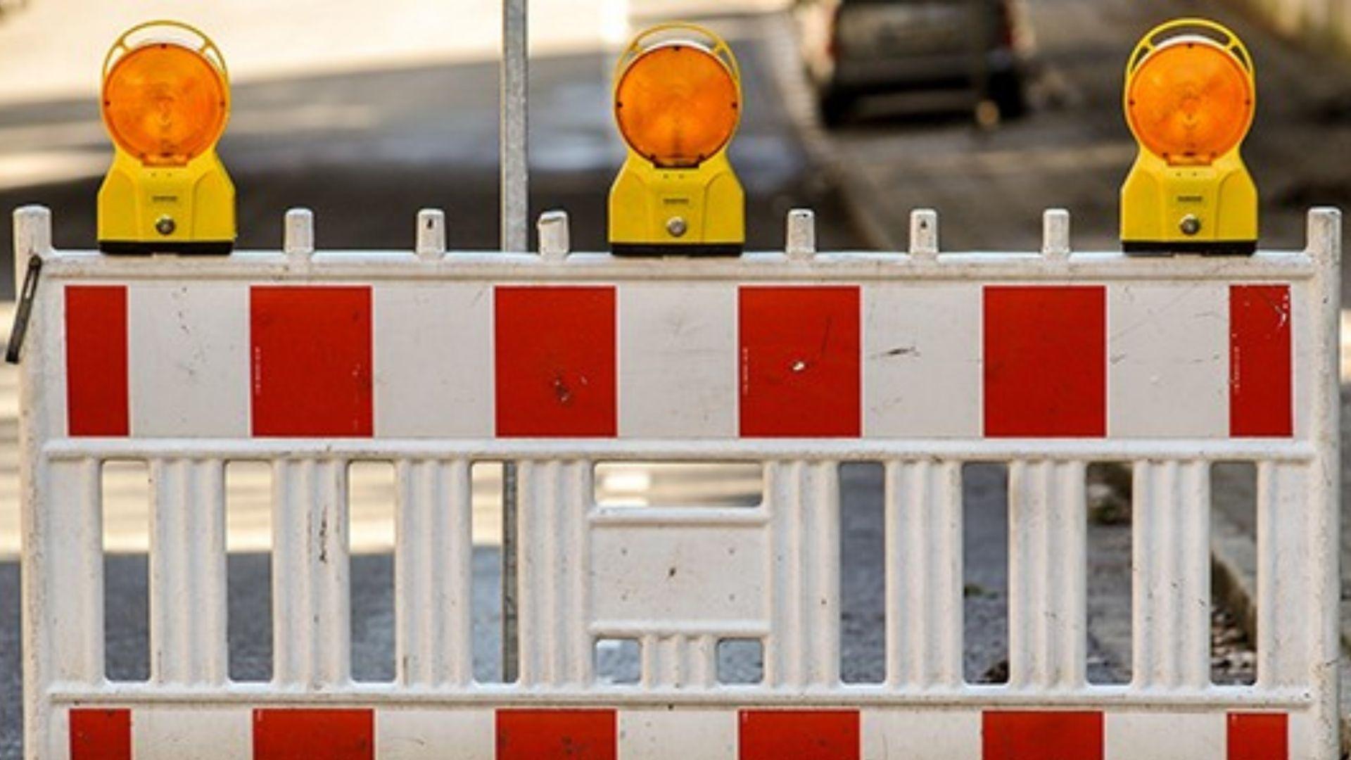 Abgebildet ist ein sogenannter rot-weißer Schrankenzaun, mit dem Baustellen oder Durchfahrten abgesperrt werden. Auf dem Zaunelement sind drei orangefarbene Warnleuchten angebracht.