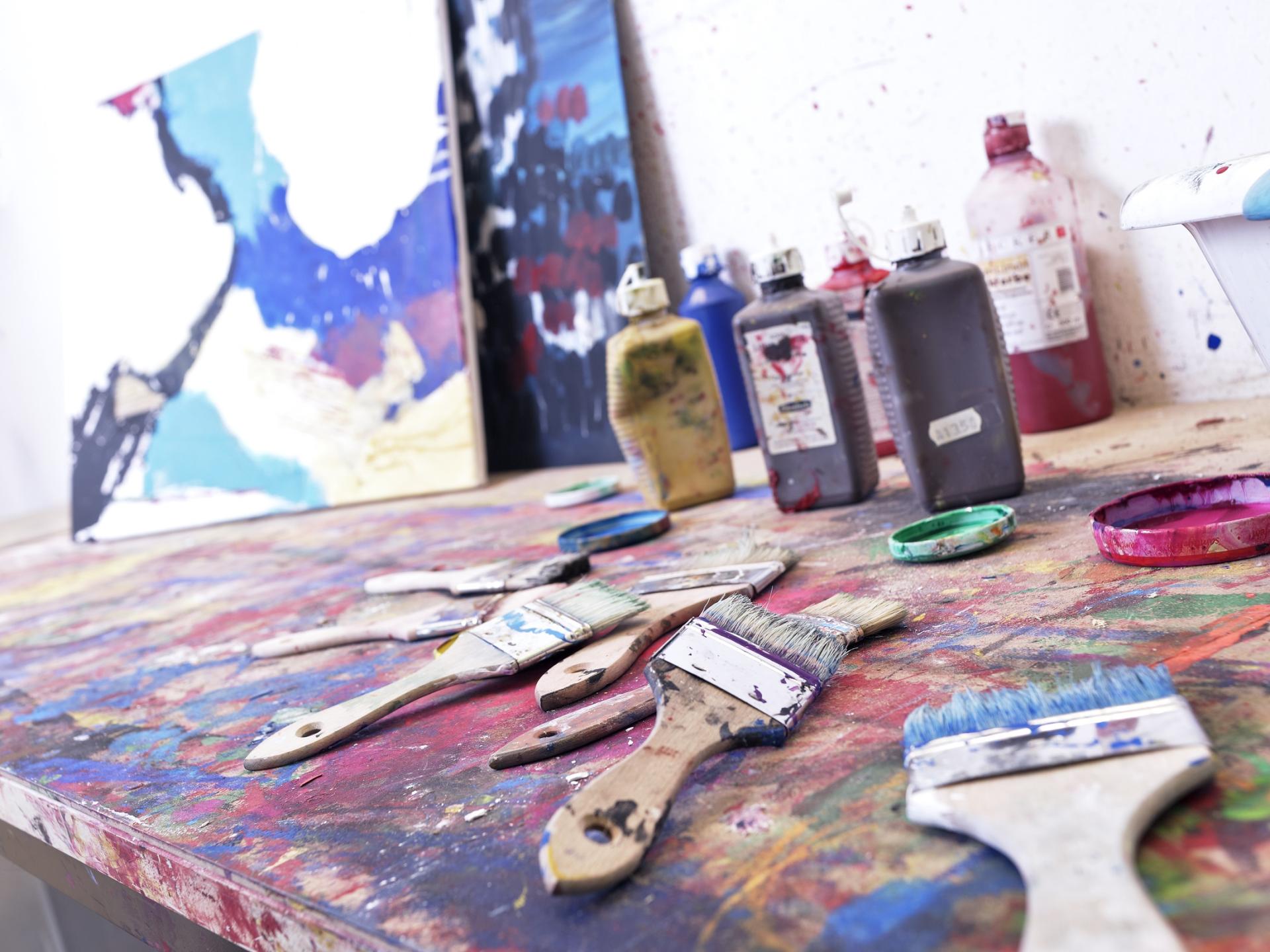 Das Bild zeigt Pinsel und Farben, im Hintergrund ist eine teilweise bemalte Leinwand zu erkennen. Die Tische sind mit Farbklecksen übersät.