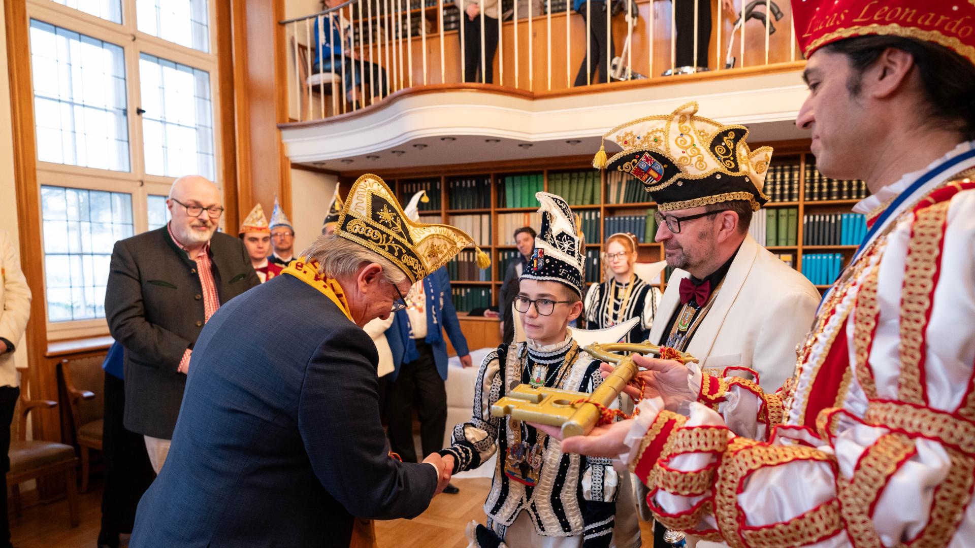 Mehrere Personen in festlichen Narrenkostümen übergeben in einem Raum mit großen Fenstern und Galerie einen goldenen Schlüssel.