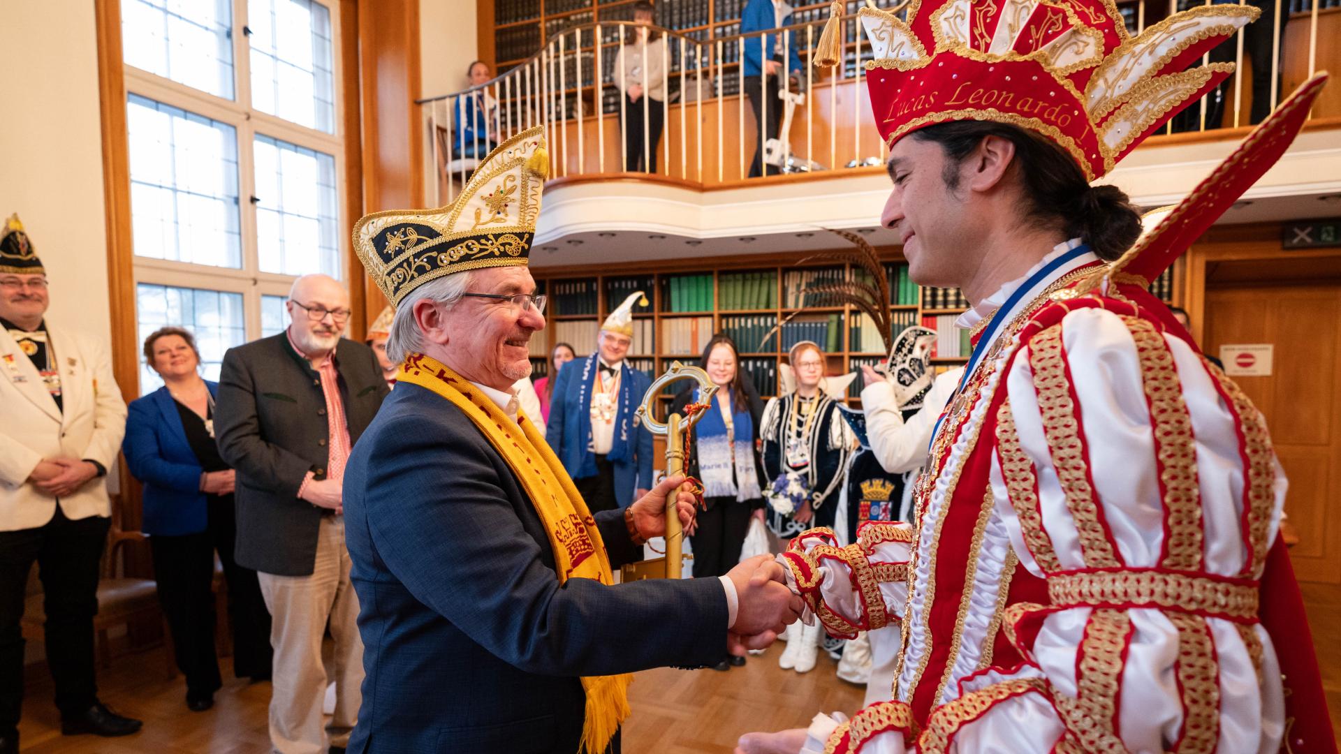 Zwei Männer in festlicher Kleidung und Narrenkappen geben sich in einem Saal die Hand, im Hintergrund stehen weitere kostümierte Personen.