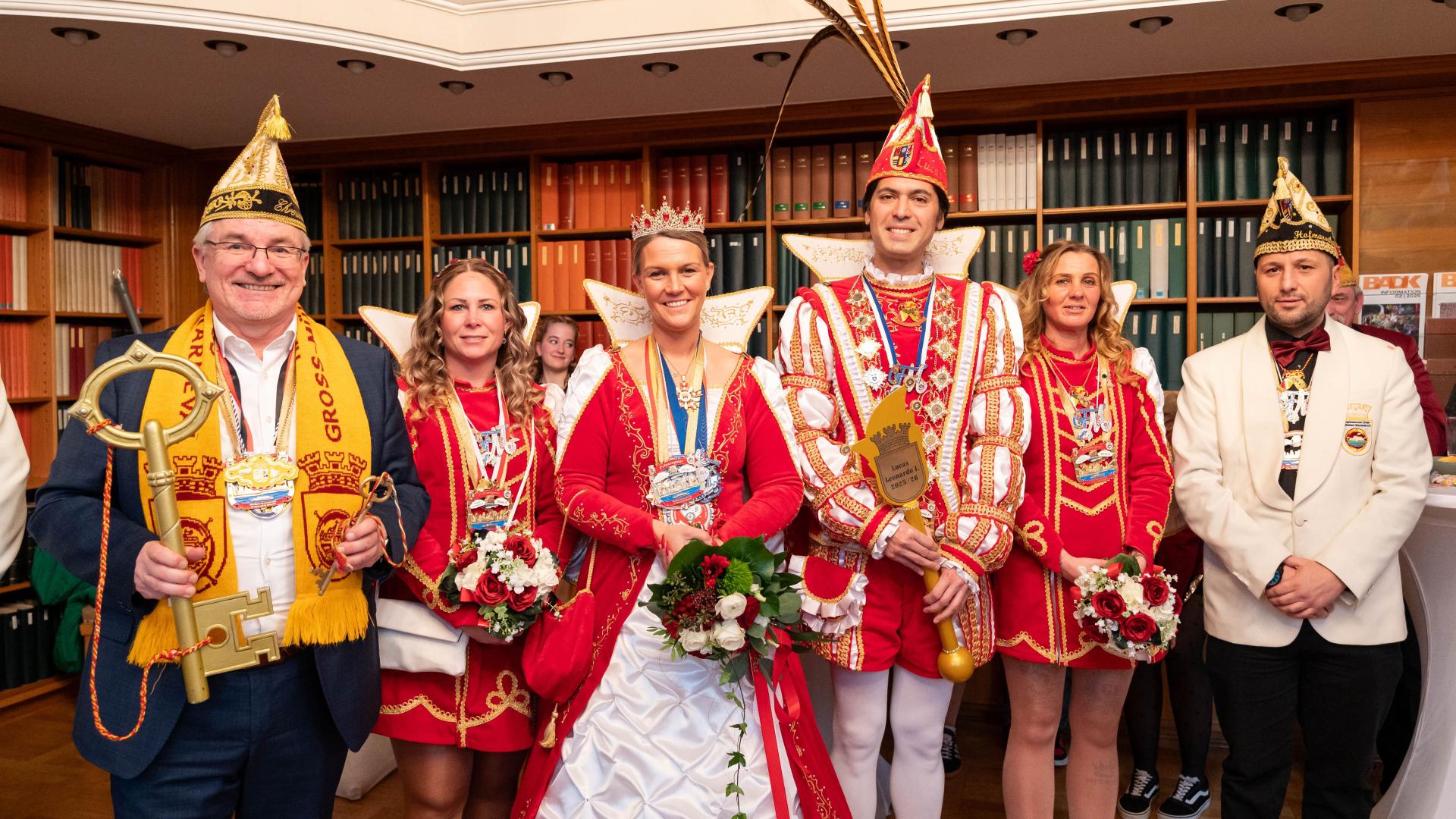 Sechs Personen in festlicher Karnevalskleidung stehen vor einem Bücherregal, darunter zwei in rot-weißen Prinzenkostümen mit Kronen und vier weitere in bunten Kostümen, Oberbürgermeister Marc Buchholz hält die goldenen Schlüssel.