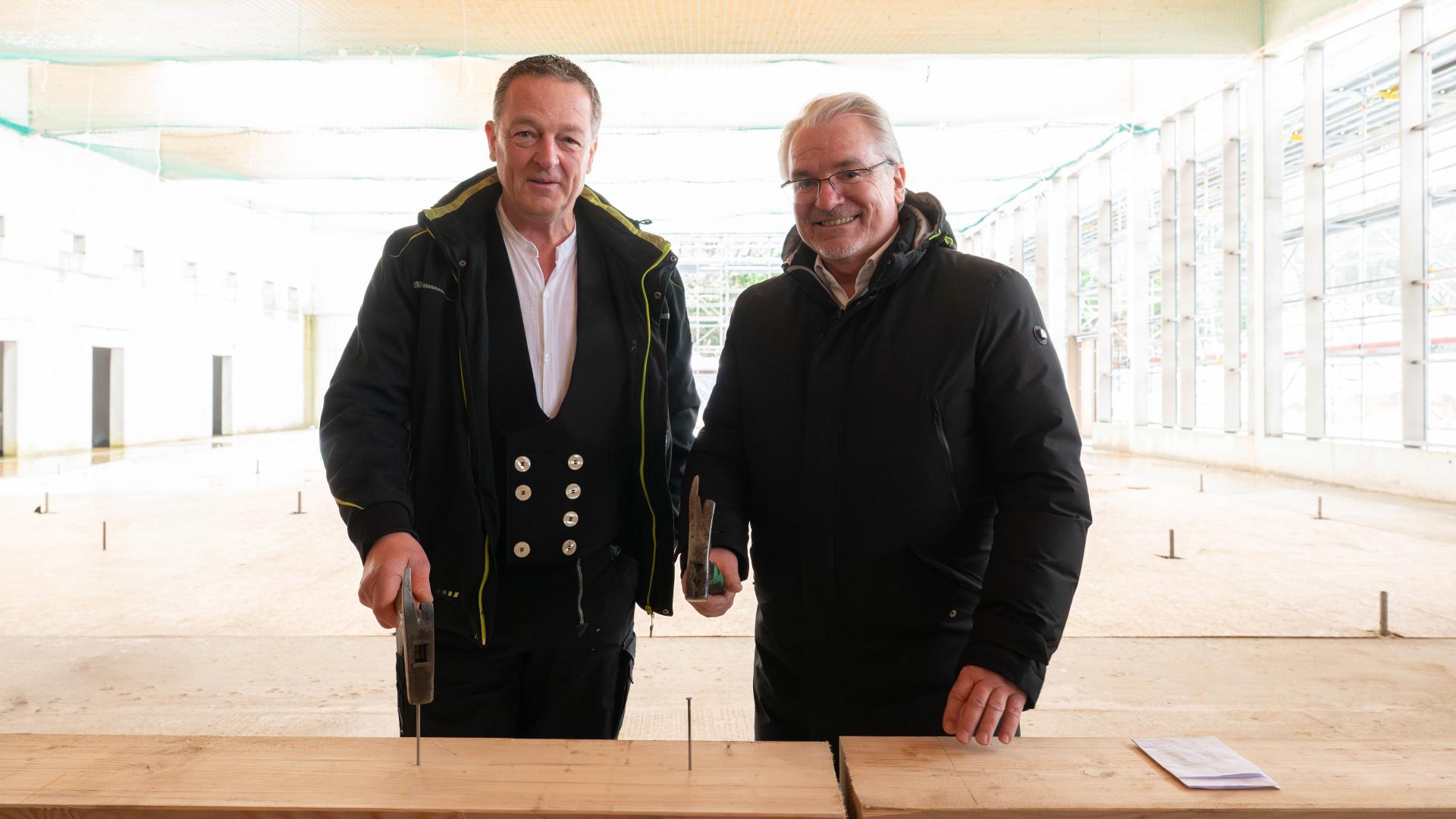 Zwei Männer in schwarzer Arbeitskleidung stehen in einer großen Schwimmhalle vor einem Holzbrett mit zwei Nägeln darin, beide halten einen Hammer.