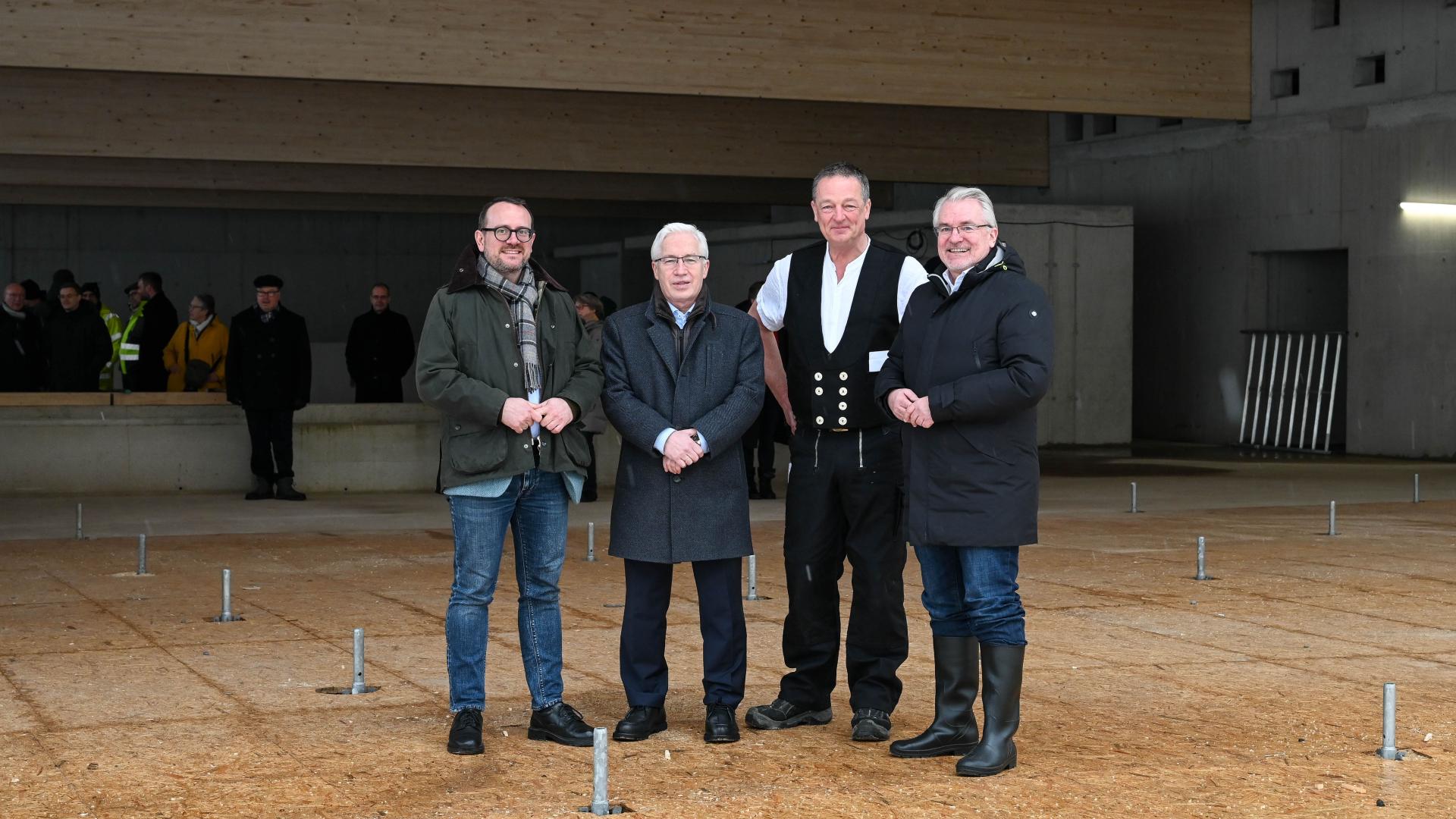 Vier Männer stehen in einer großen, halbfertigen Schwimmhalle. Auf dem Boden sind Metallstangen, im Hintergrund weitere Personen und eine Holzdecke