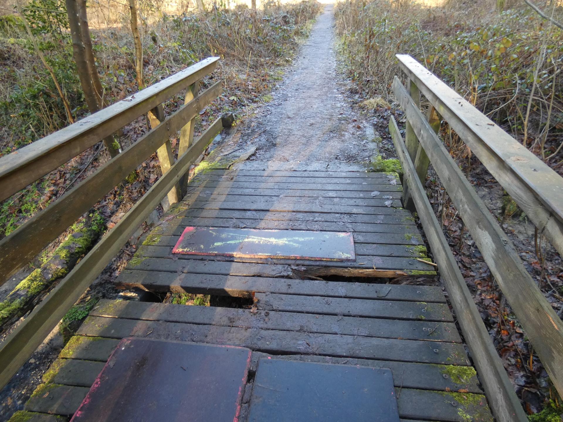 Abgebildet ist eine Holzbrücke mit Löchern in den Bodenplanken.