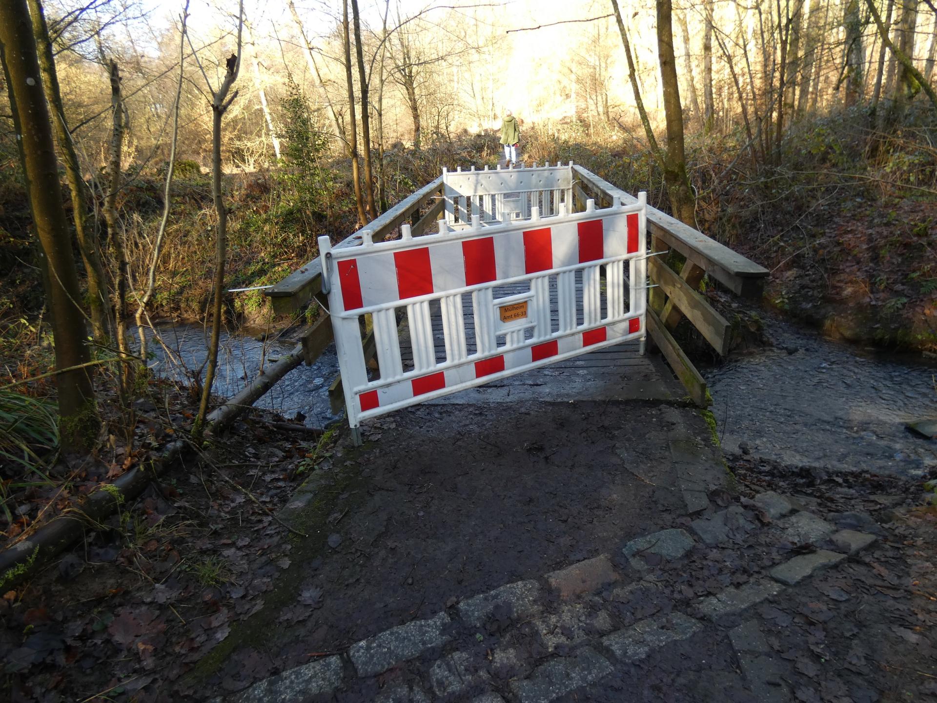 Abgebildet ist eine mit rot-weiß gestreiften Baken abgesperrte Holzbrücke für Fußgänger im Wald des Rumbachtals.