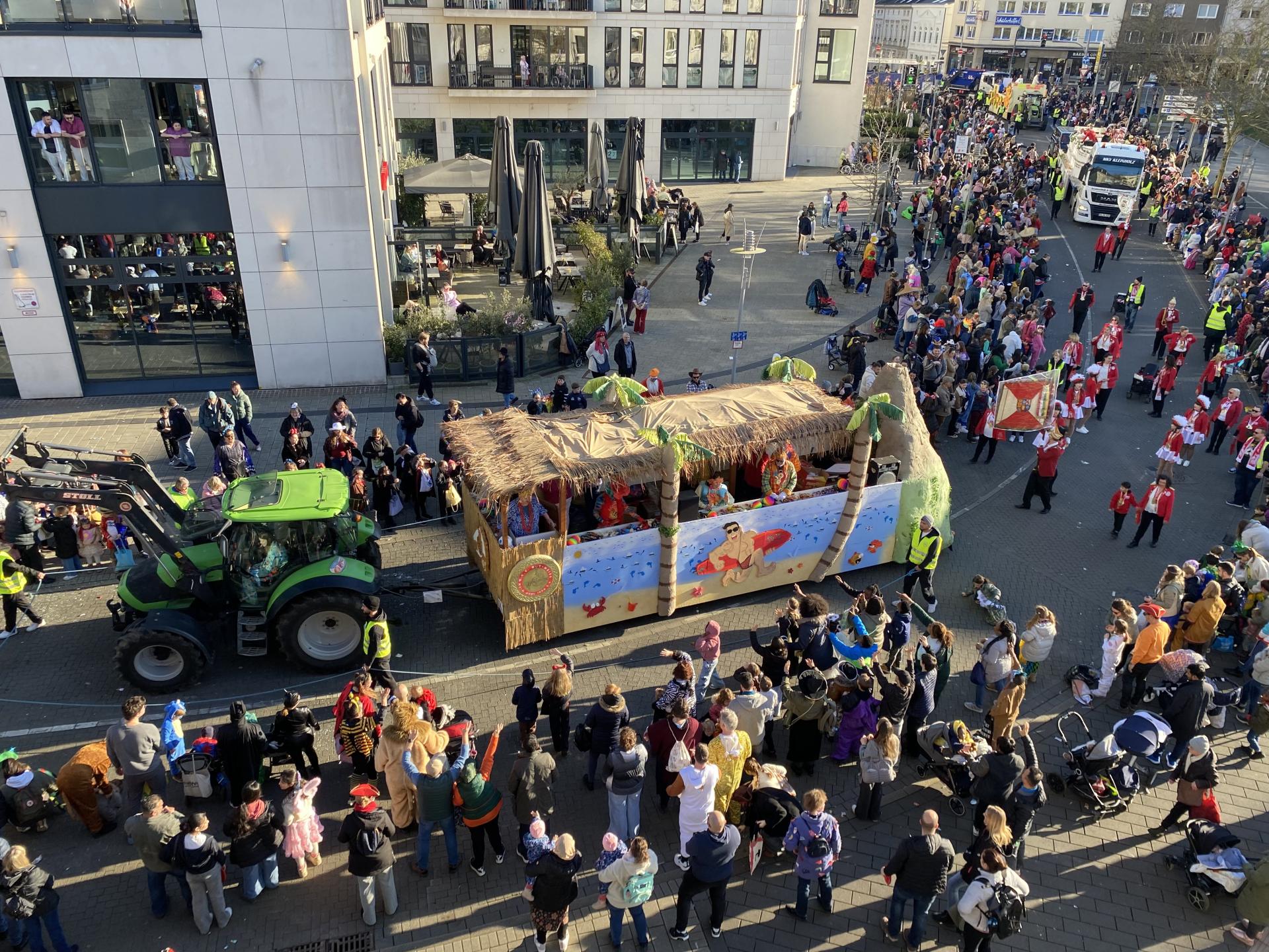 Blick aus dem Rathaus auf den Rosenmontagszug in der Schollenstraße. Ein Trecker zieht einen Mottowagen. Im Hintergrund folgen Fußgruppen und weitere Mottowagen des Mülheimer Umzugs.
