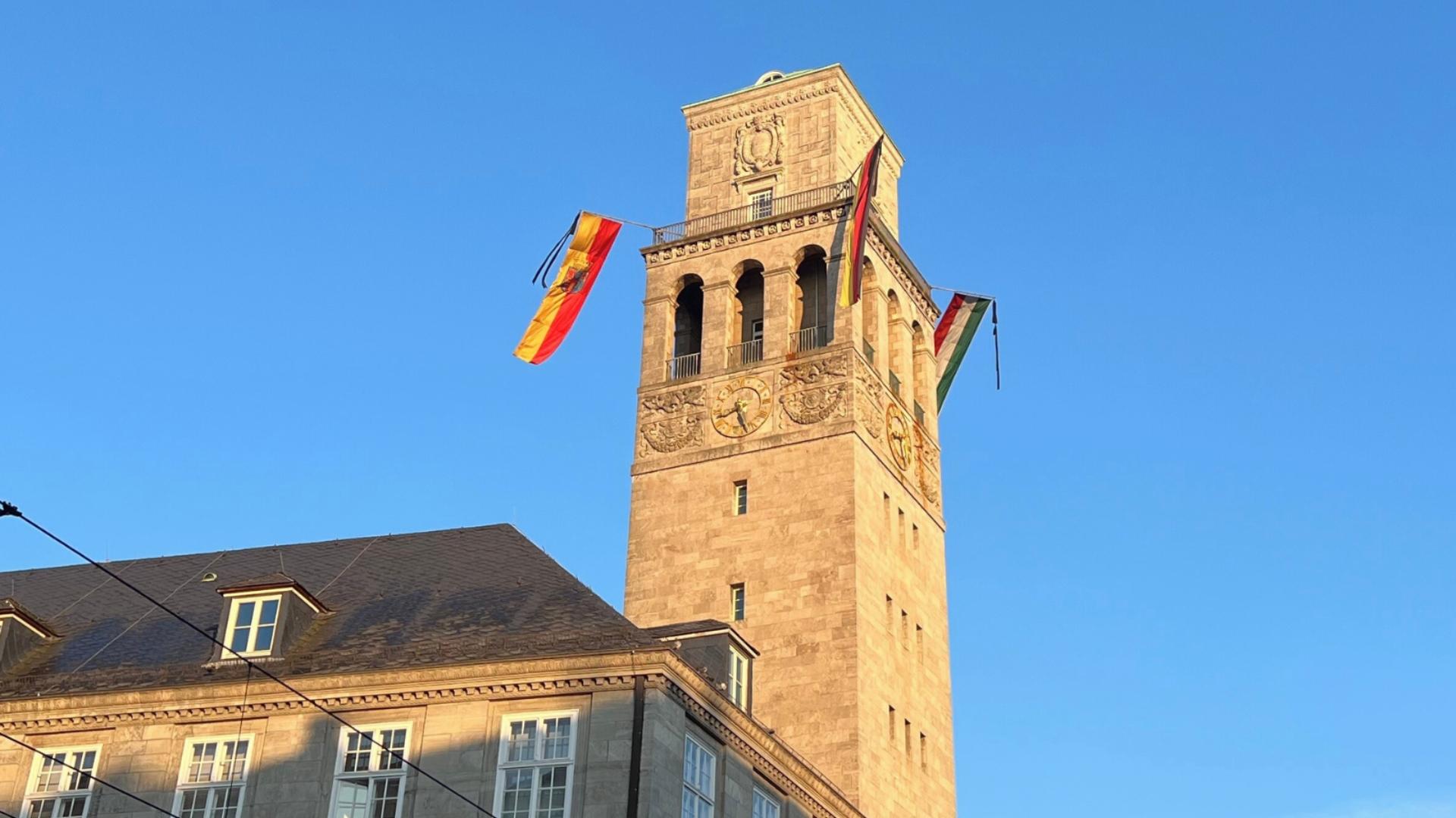 Foto zeigt die Trauerbeflaggung des Mülheimer Rathausturmes. Die Trauerbeflaggung besteht aus den Flaggen der Stadt Mülheim an der Ruhr, des Landes Nordrhein-Westfalen, von Deutschland und Europa sowie jeweils einen Trauerflor.