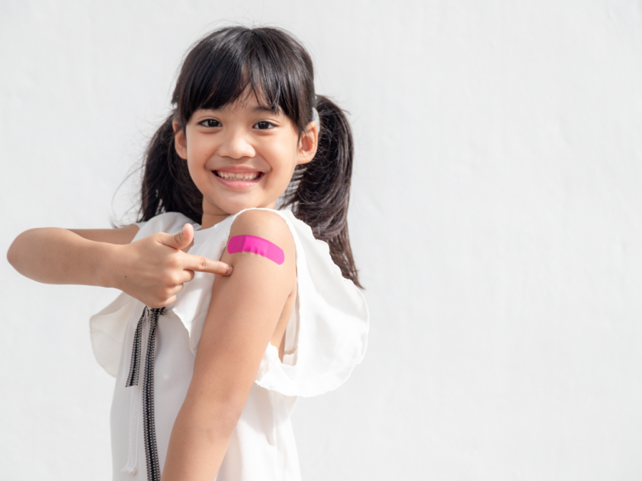 Mädchen mit zwei Zöpfen zeigt lächelnd auf ihren Arm, auf dem ein pinkes Pflaster nach der Impfung klebt.