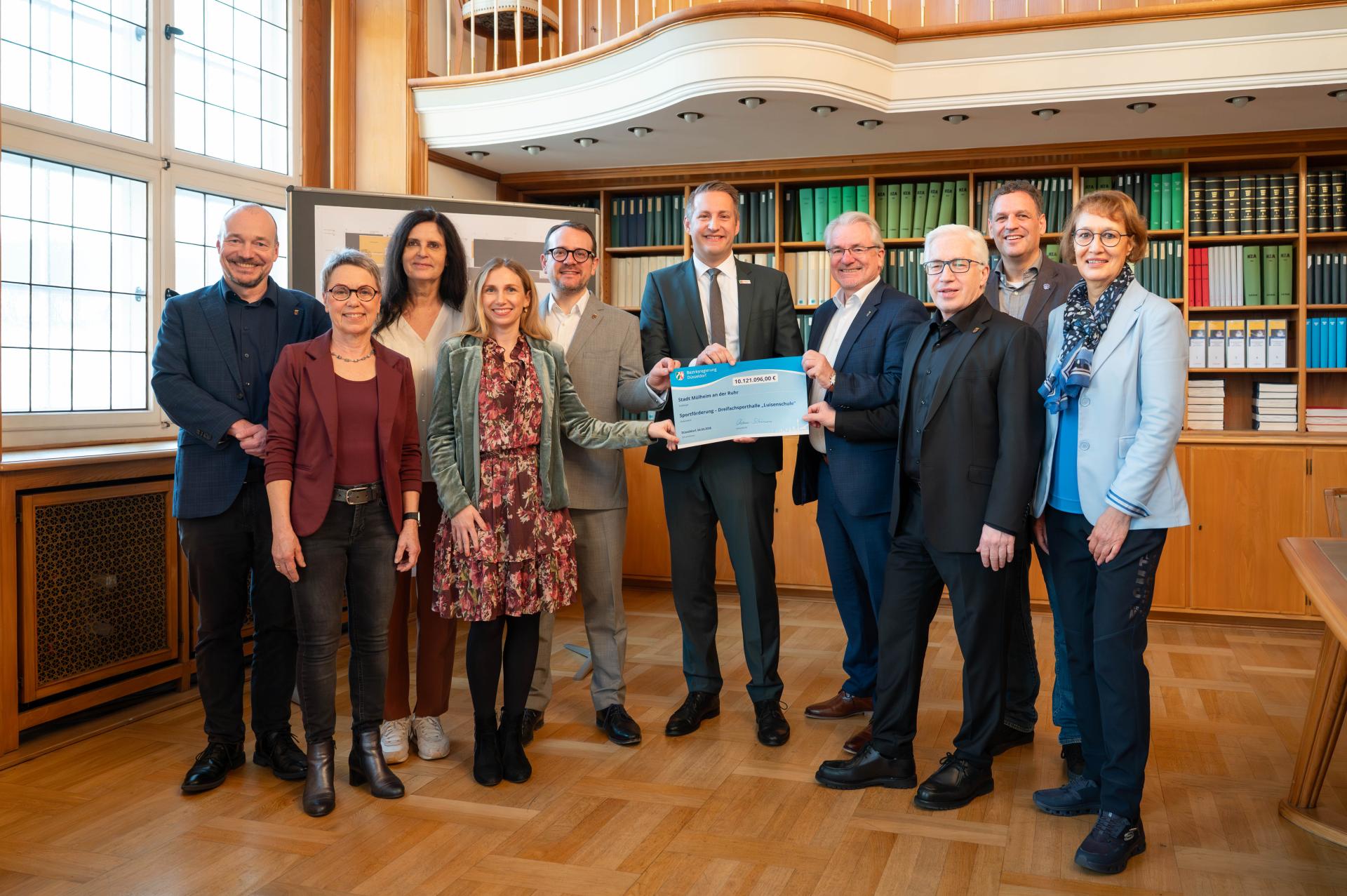 Zehn Personen stehen in einem Raum mit Holzregalen und halten gemeinsam einen großen blauen Förderbescheid für die Dreifachsporthalle der Luisenschule. Rathausbibliothek