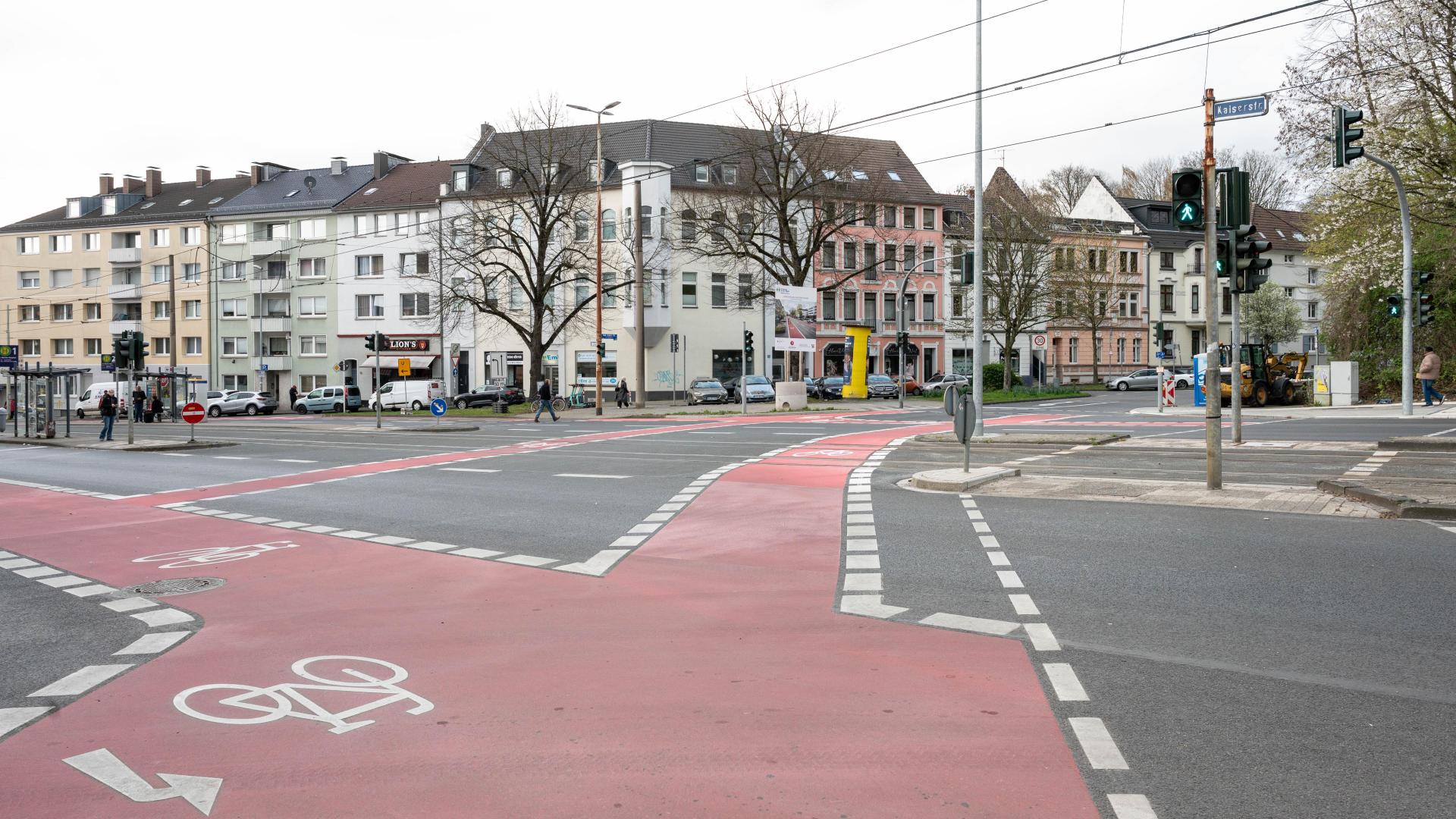 Breiter roter Radweg mit Fahrradpiktogrammen an einer Kreuzung vor mehrstöckigen Wohngebäuden und Fußgängern