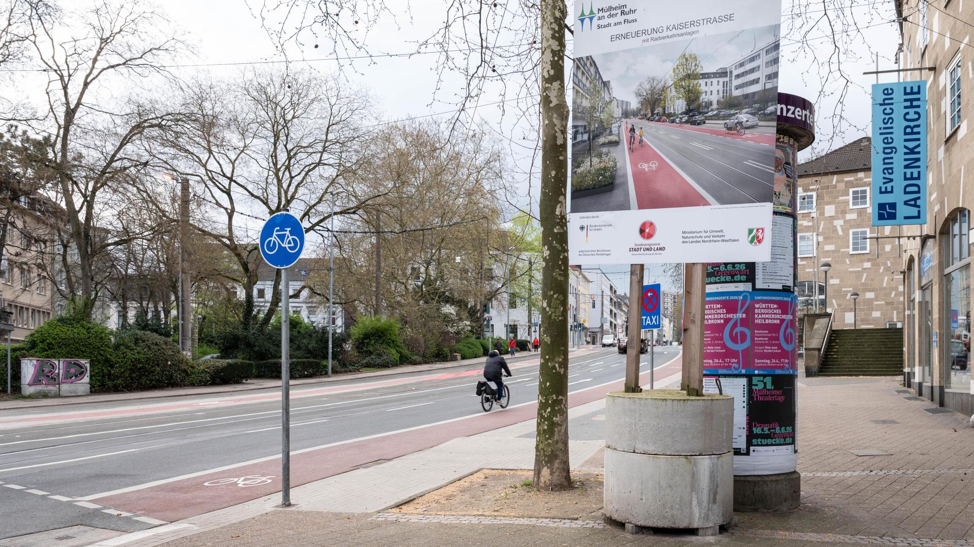 Straße mit neuem roten Radweg, Fahrrad-Verkehrsschild und Plakat zur Erneuerung der Kaiserstraße neben einem Baum und einer Litfaßsäule